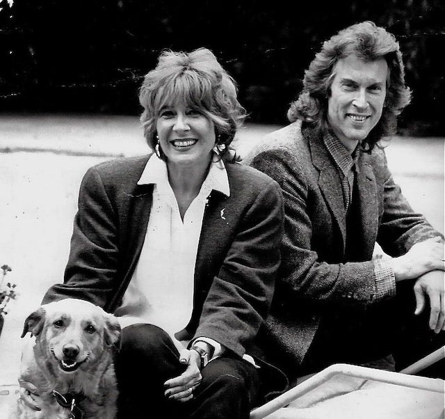 A woman and a man sit outdoors, both smiling, with a dog sitting beside the woman. All are dressed in casual or semi-formal clothing. The background is blurred.