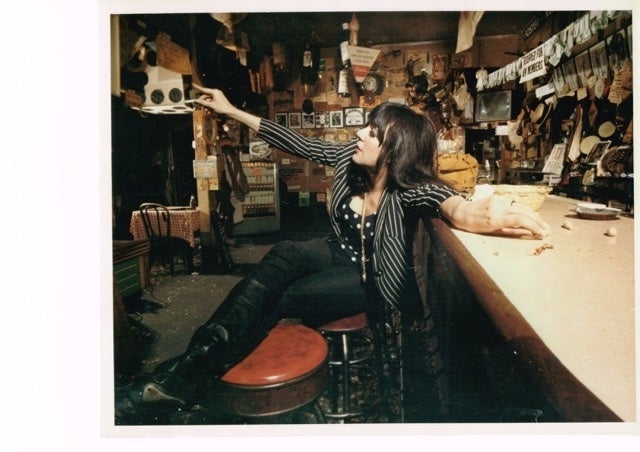 A person sits on a barstool at a cluttered bar, leaning back and pointing upwards, with various objects and decorations in the background.