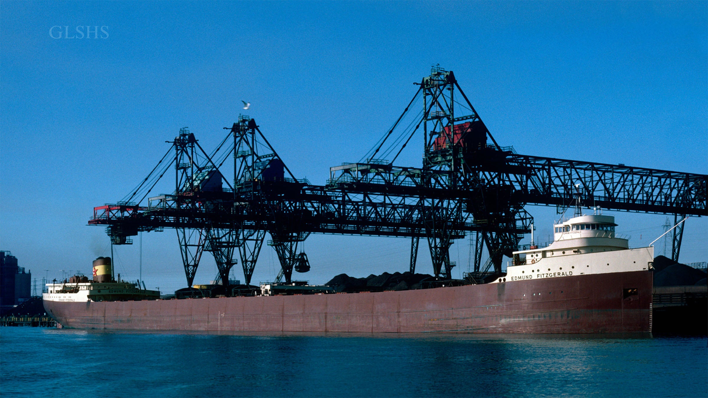 A large cargo ship is docked at an industrial port facility, with cranes positioned above to load or unload materials.