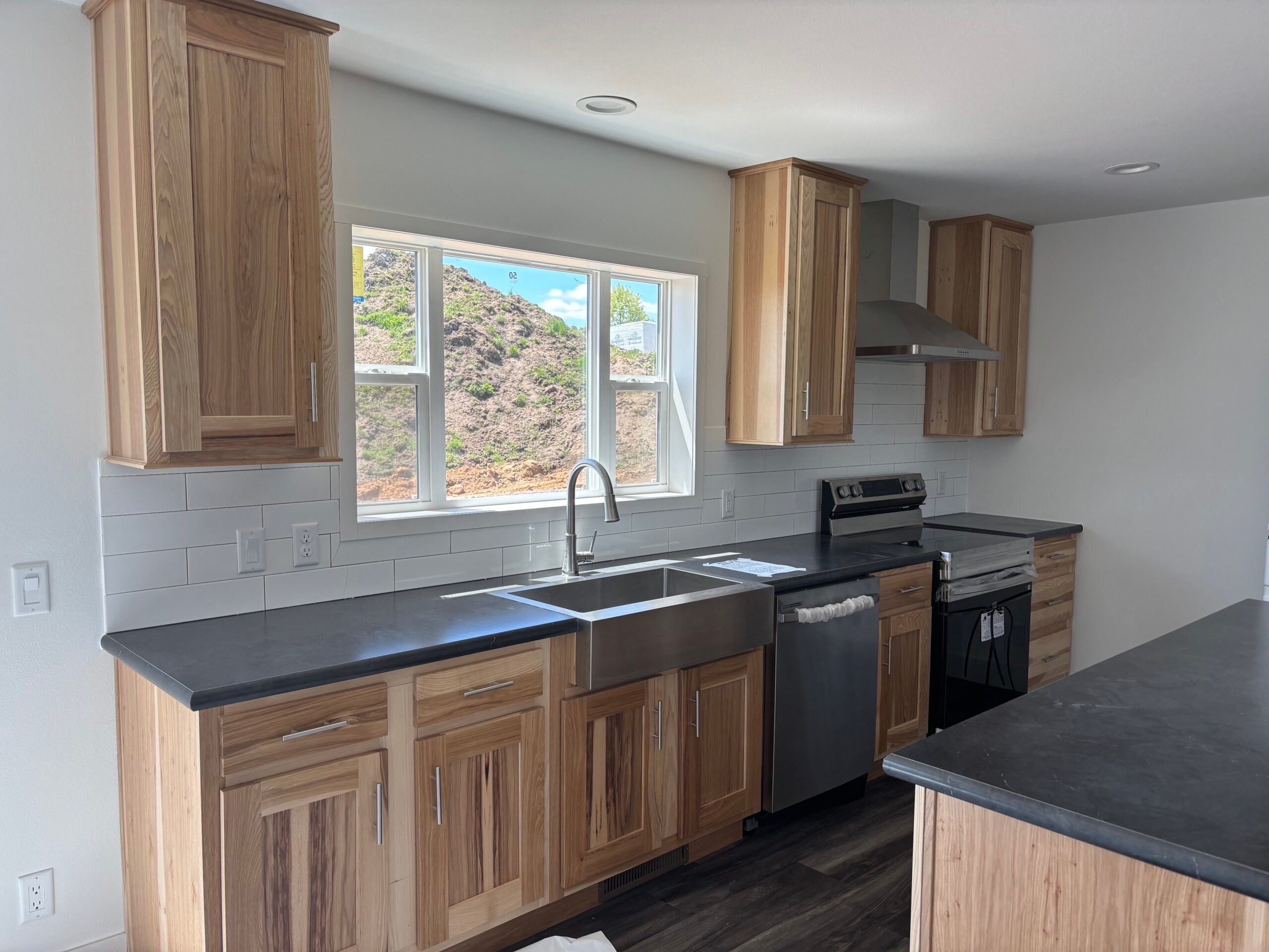 Modern kitchen with wooden cabinets, stainless steel appliances, farmhouse sink, and a large window above the sink, showing a hillside view outside.