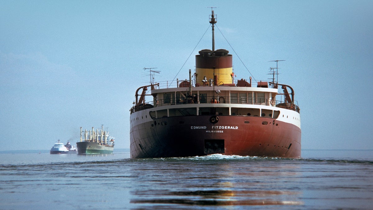 A large cargo ship named Edmund Fitzgerald is sailing on calm water, with two other ships visible in the distance under a clear sky.