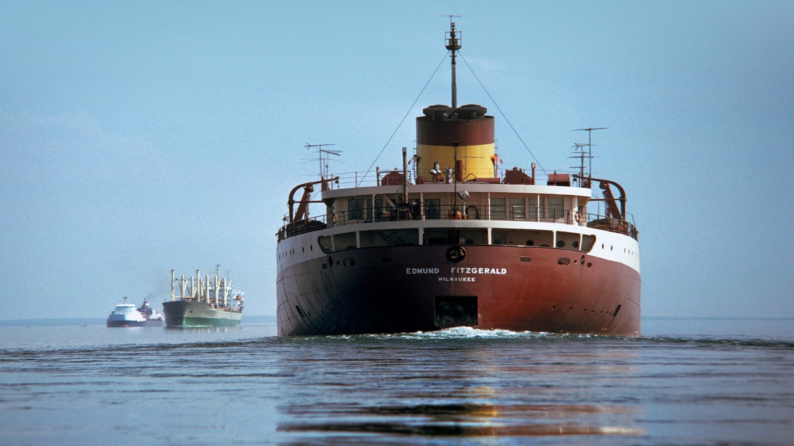 A large cargo ship named Edmund Fitzgerald is sailing on calm water, with two other ships visible in the distance under a clear sky.