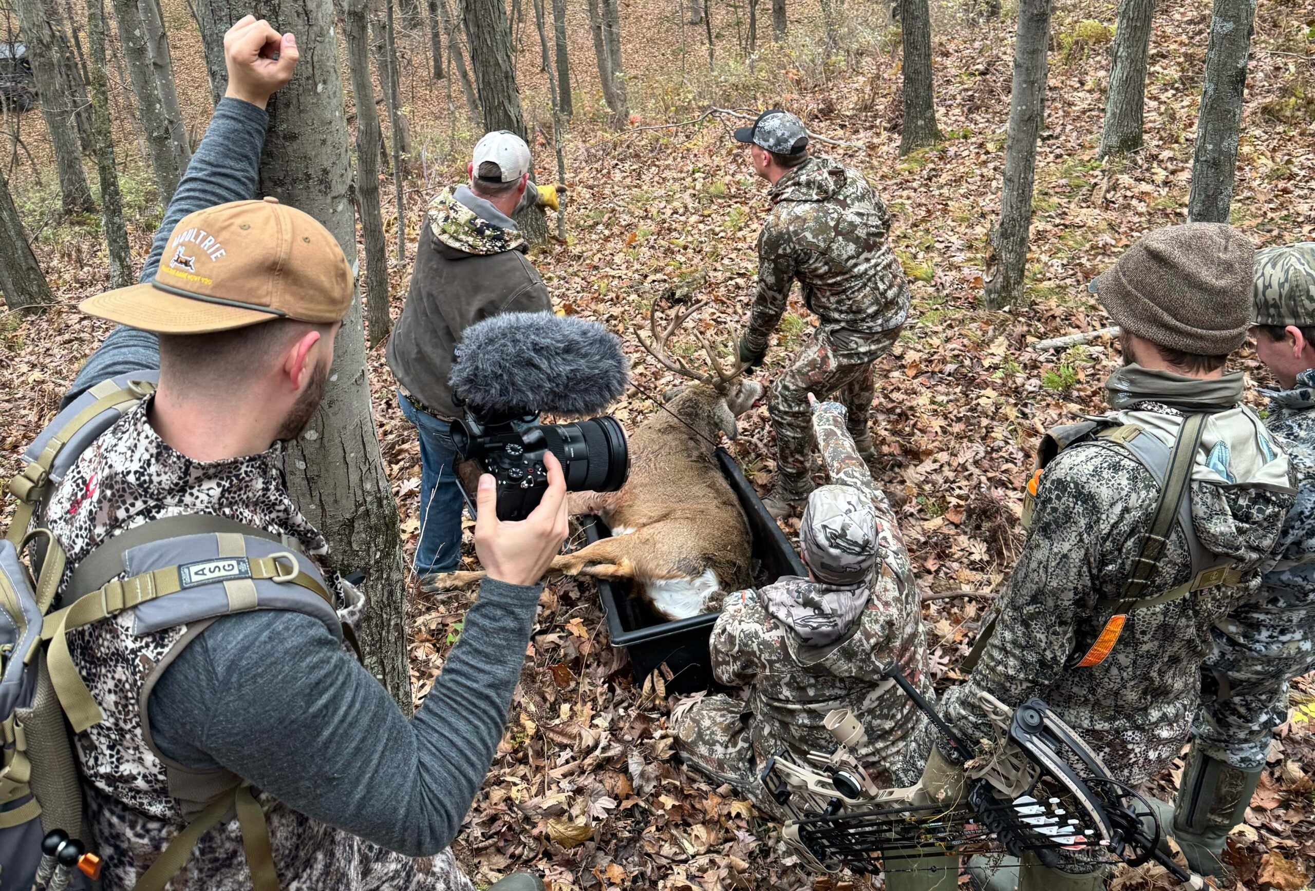 Five hunters wearing camouflage stand in a forest around a large deer on the ground; one holds a camera, and fallen leaves cover the forest floor.