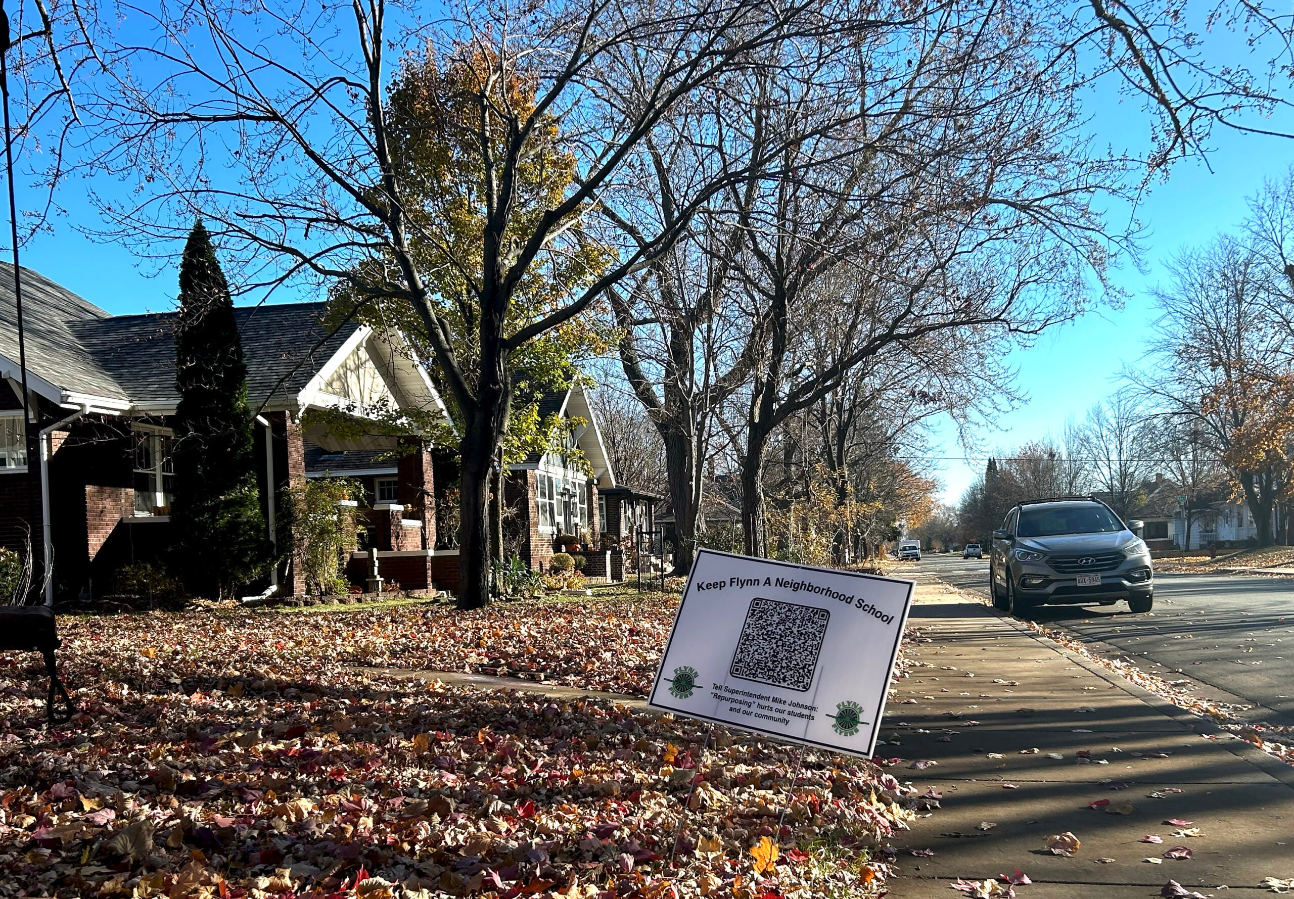 A tree-lined residential street with autumn leaves on the ground and a sign on the sidewalk displaying a QR code and the message, Keep Flynn A Neighborhood School.