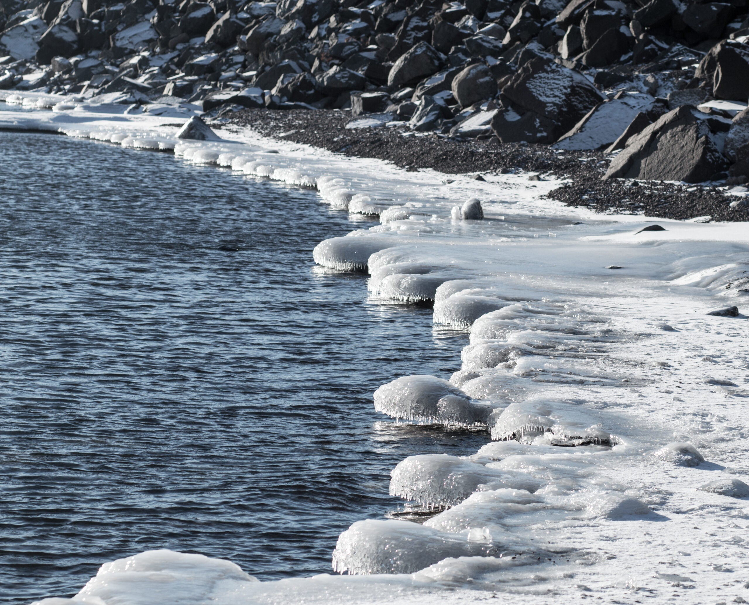 A rocky shoreline with partially frozen water and snow-covered ice formations along the edge.
