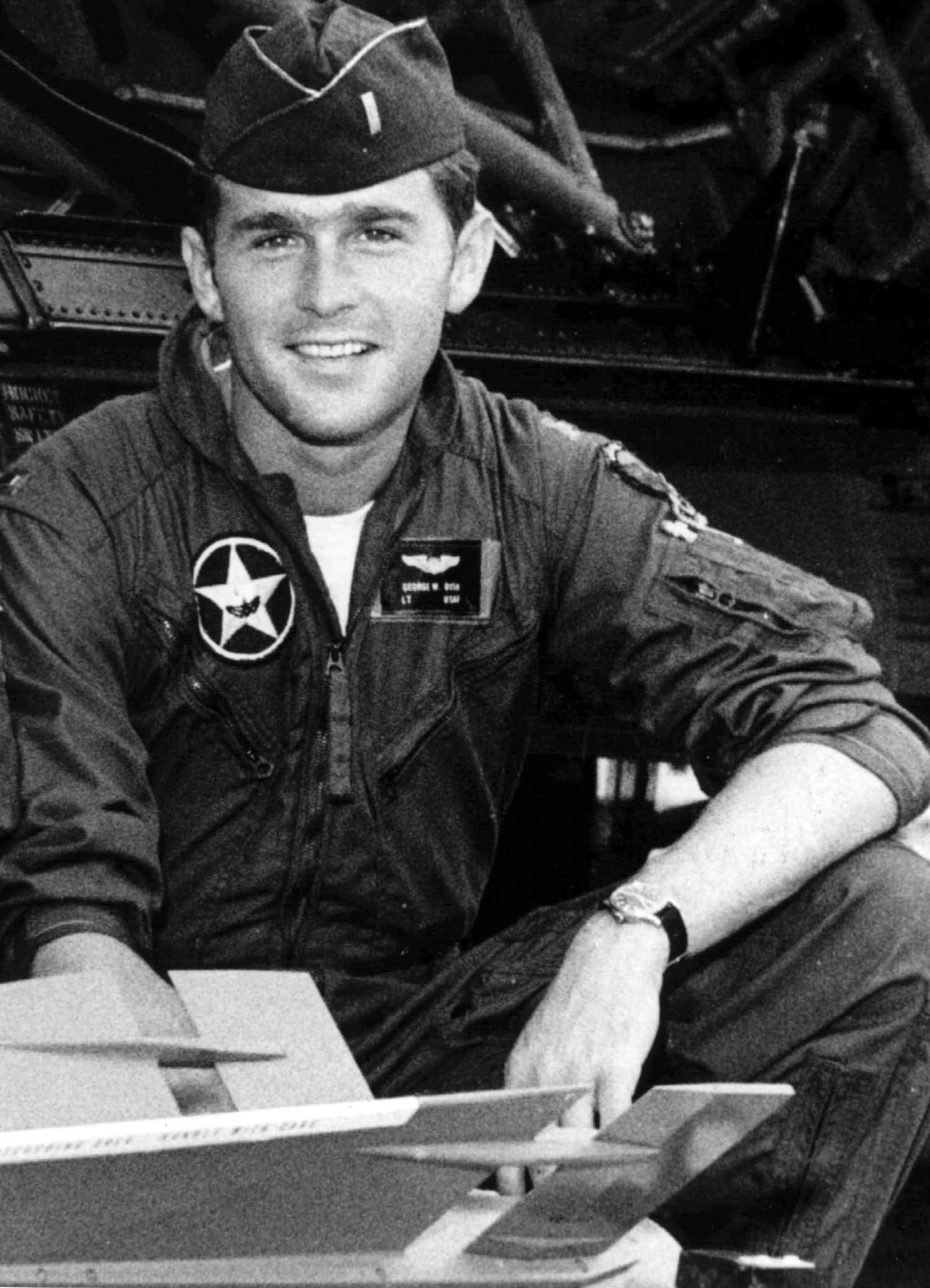 A man in a military flight suit and cap sits near aircraft equipment, smiling at the camera.