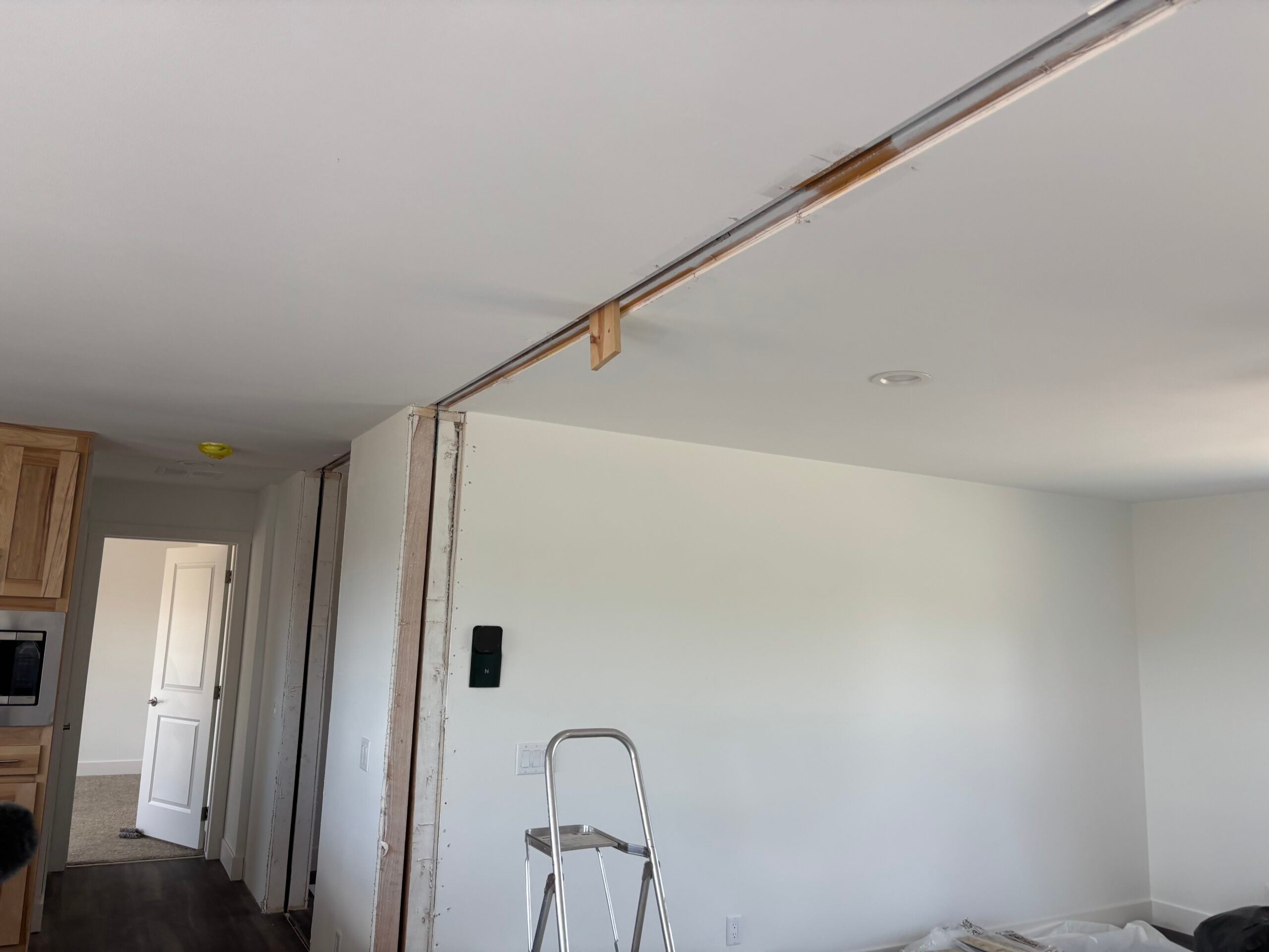 A partially removed interior wall with exposed studs and a support beam along the ceiling; a ladder and remodeling materials are visible in the room.