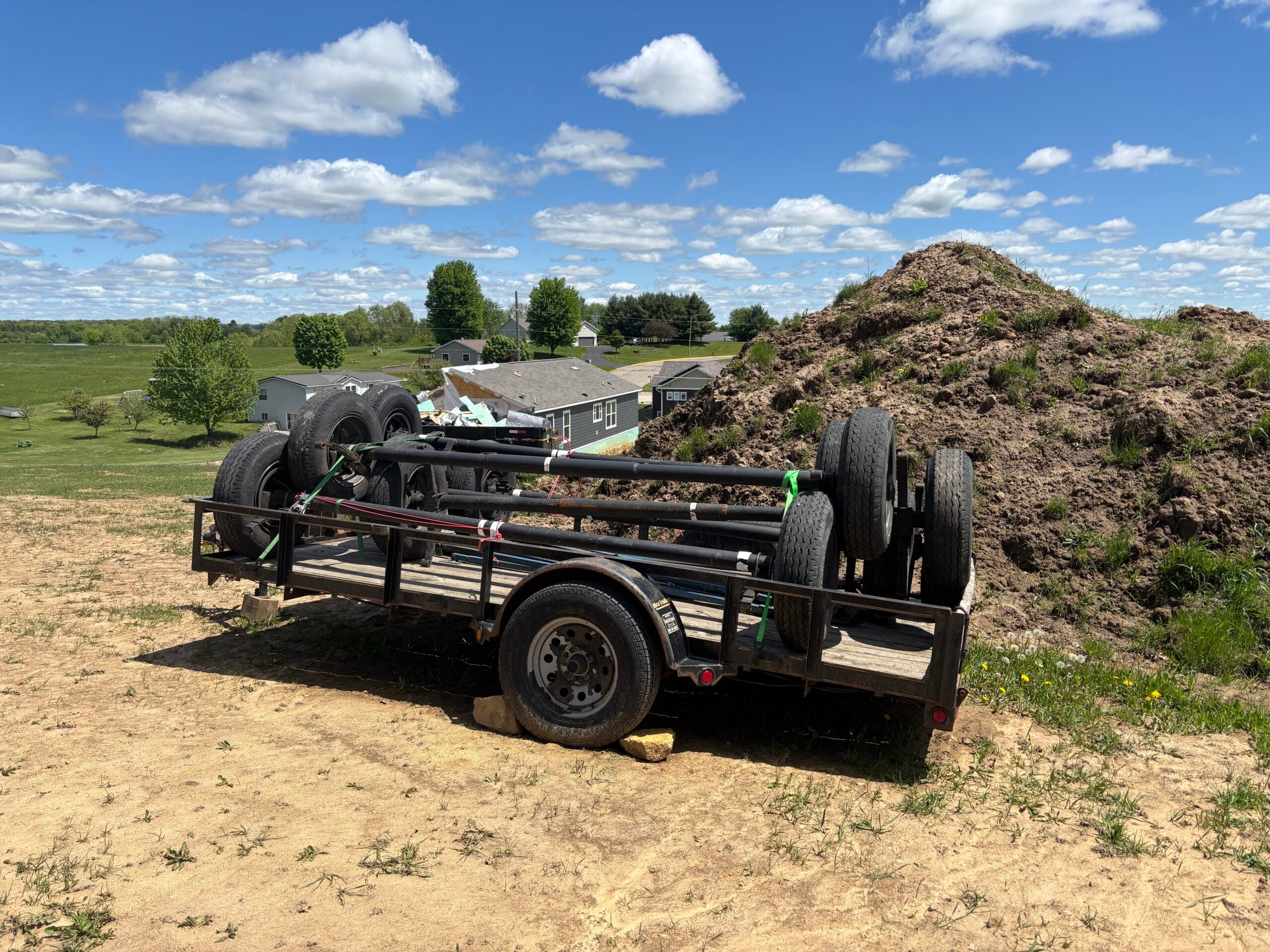 A black utility trailer loaded with metal frames and wheels sits on a dirt lot near a mound of soil, with houses and fields in the background under a partly cloudy sky.