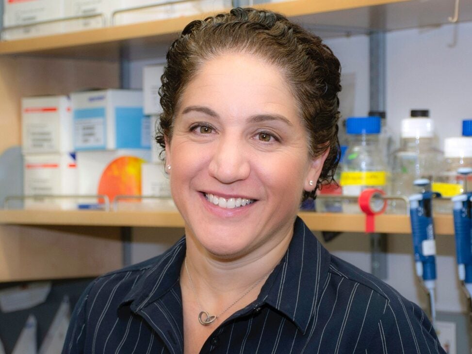 A person with short curly hair and a dark collared shirt smiles in a laboratory setting with shelves of scientific supplies in the background.
