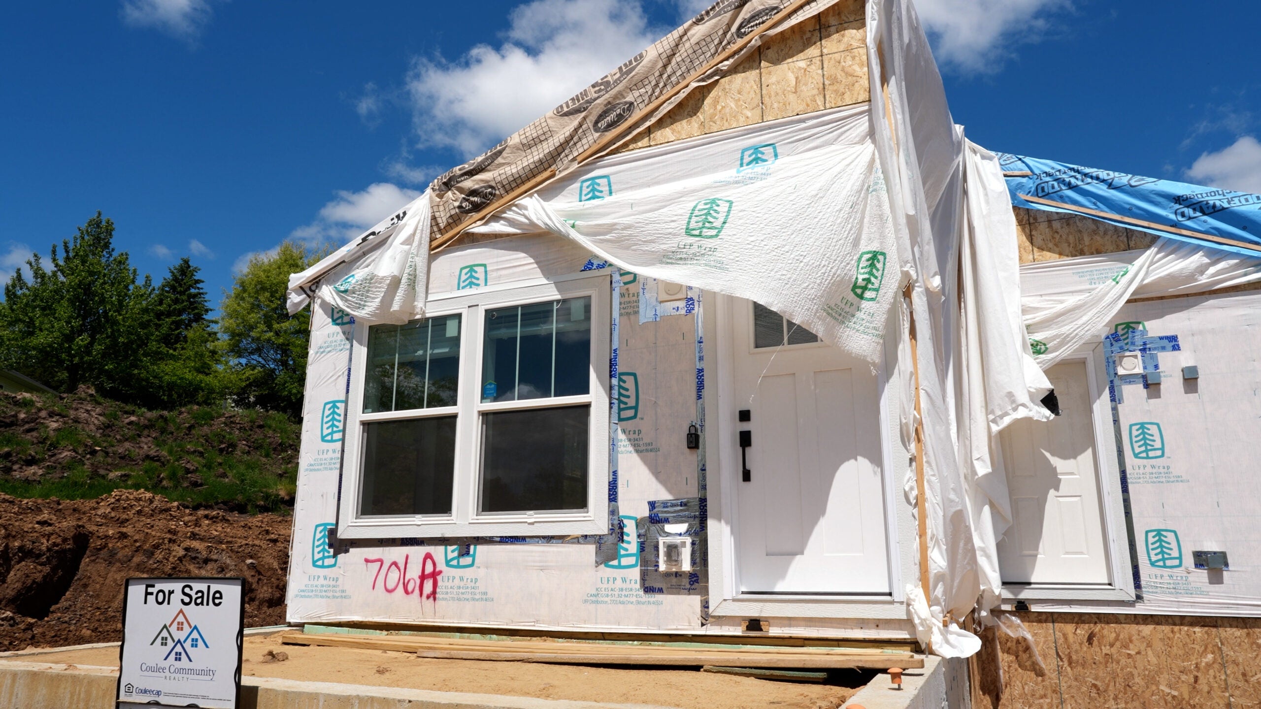 Partially constructed house with exposed insulation and tarp draped over the front; For Sale sign visible in the yard.