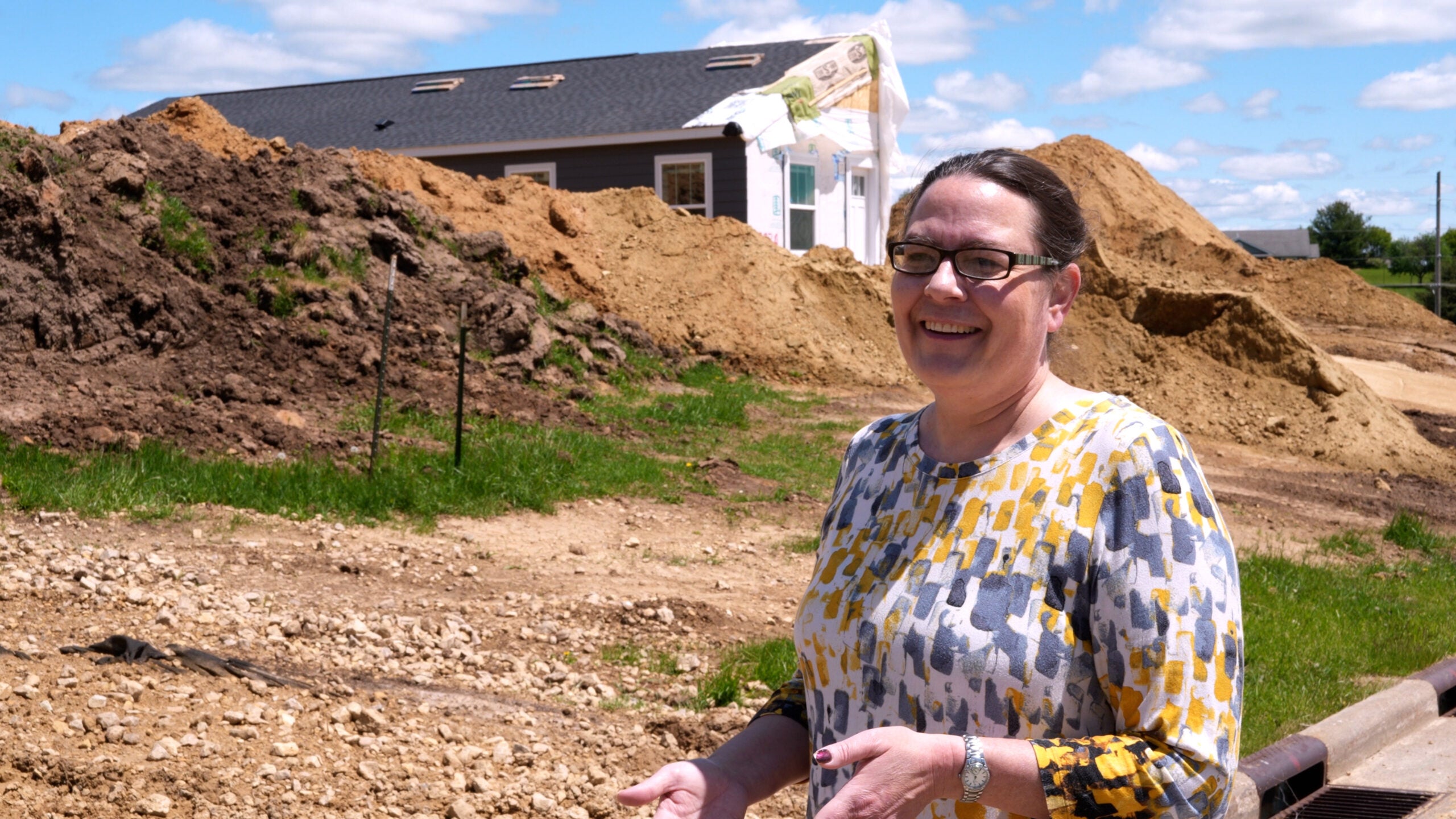 A woman in glasses stands and smiles at a construction site with piles of dirt and a partially built house in the background under a blue sky.