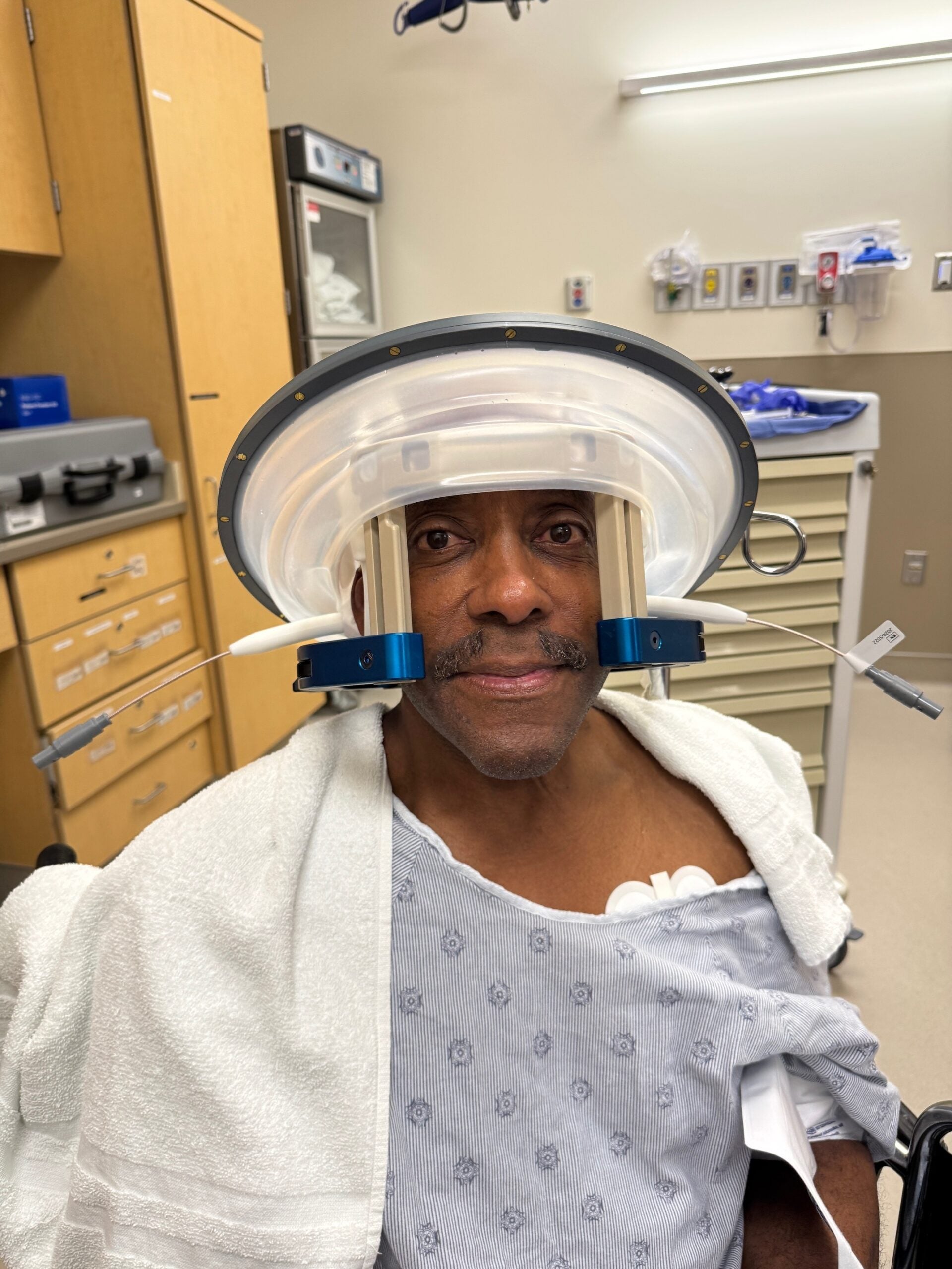 A man in a hospital gown sits in a medical room with a halo head brace device attached to his head and upper body.