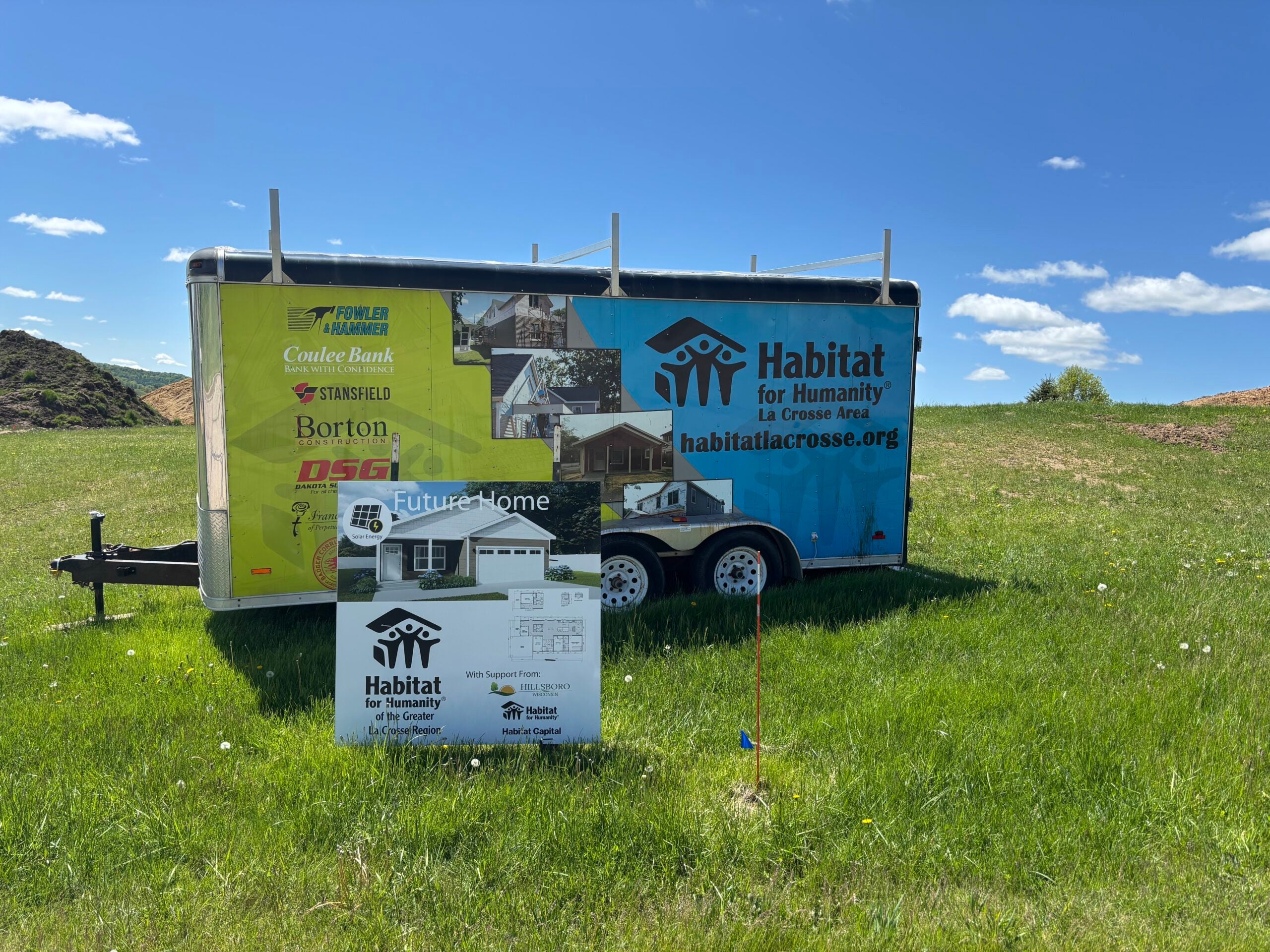 A Habitat for Humanity trailer and sign stand on a grassy field, indicating a future home construction site with sponsor logos displayed.