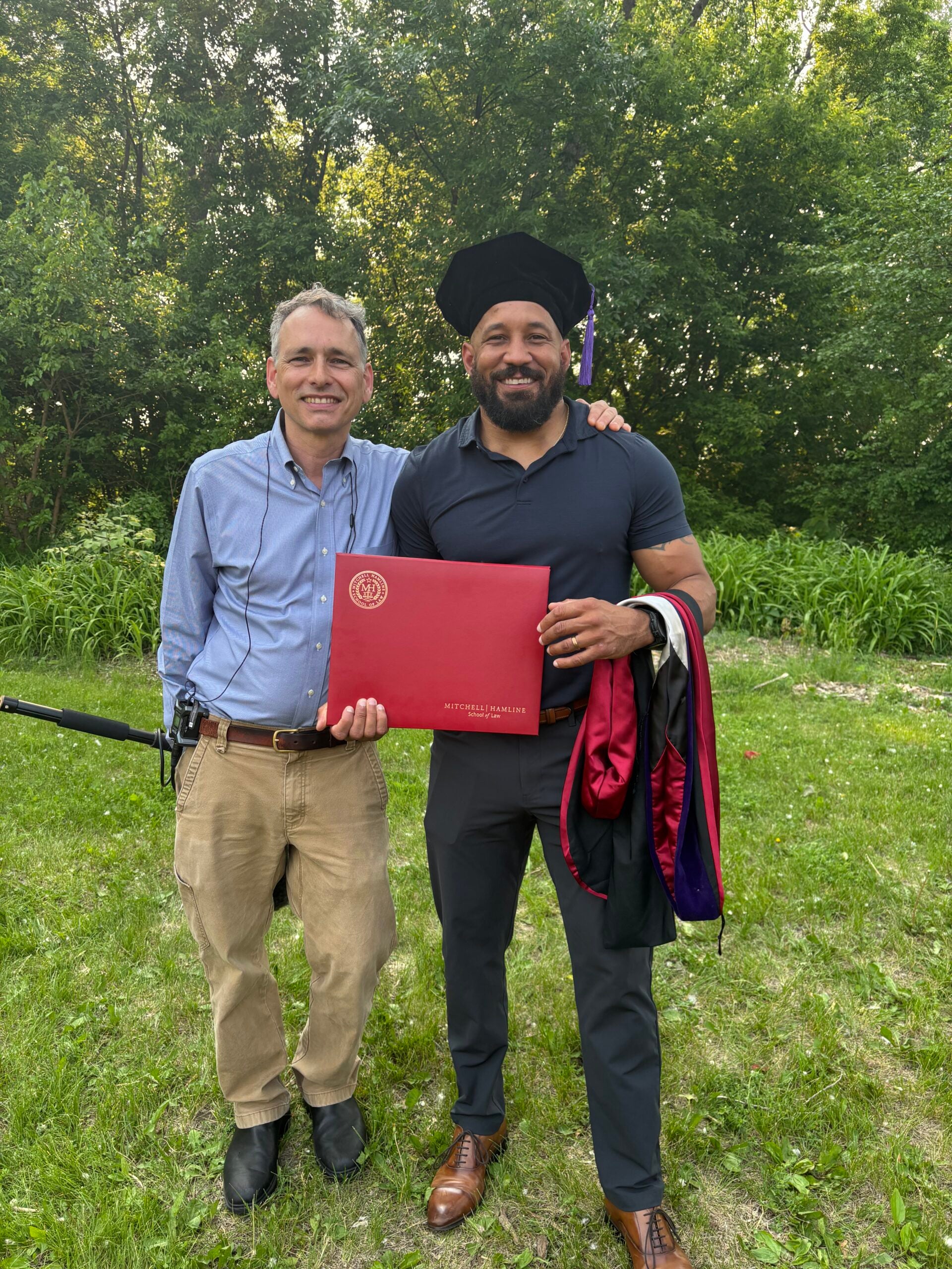 Two men stand outdoors on grass; one wears a graduation cap and holds a red diploma folder, while the other stands beside him smiling. Trees are visible in the background.