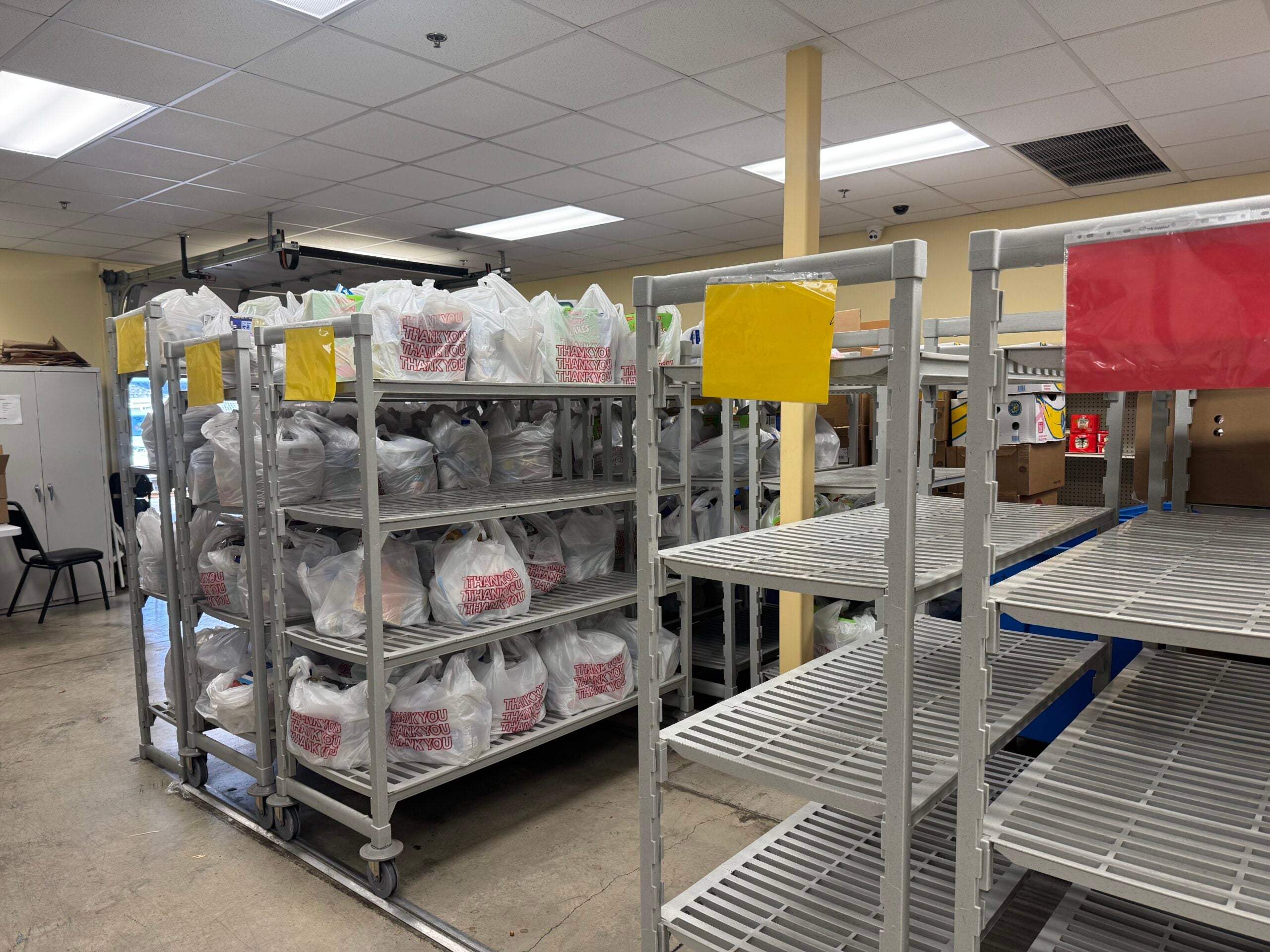 Plastic bags filled with items are stacked on metal rolling shelves, while several empty shelves are visible in the foreground of a well-lit room.