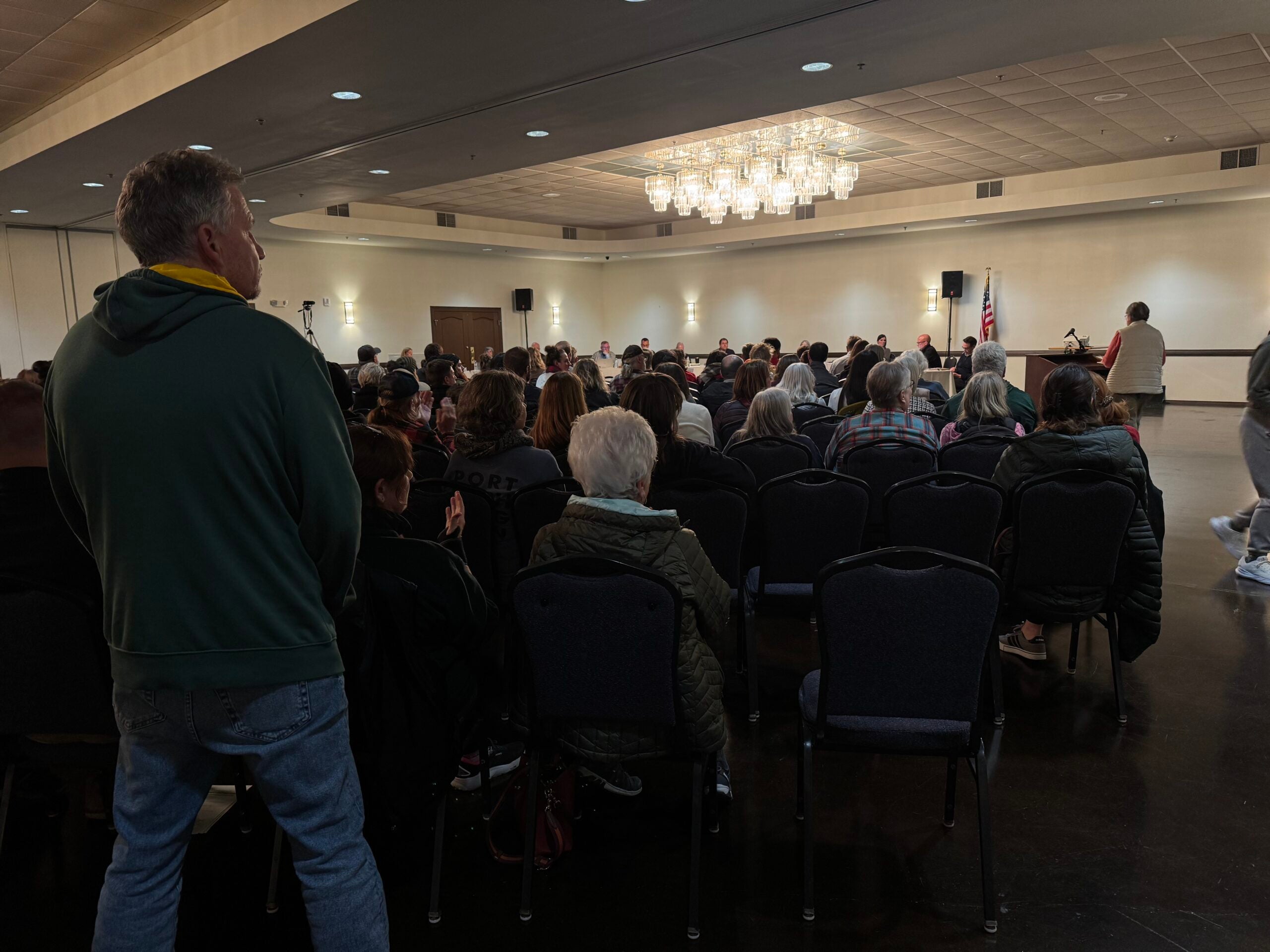 A group of people are seated in a large room with a chandelier, facing a stage area where speakers are addressing the audience; an American flag is visible.