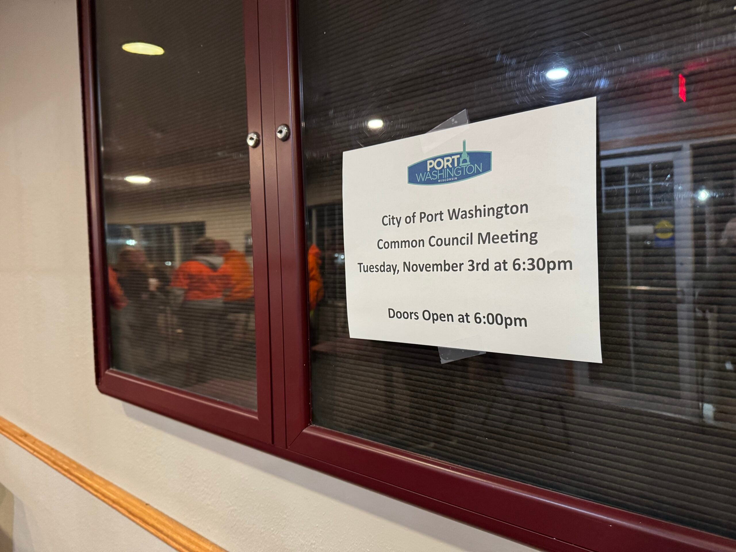 A sign on a glass window announces a City of Port Washington Common Council Meeting on Tuesday, November 3rd at 6:30pm, with doors opening at 6:00pm. People are visible in the room.