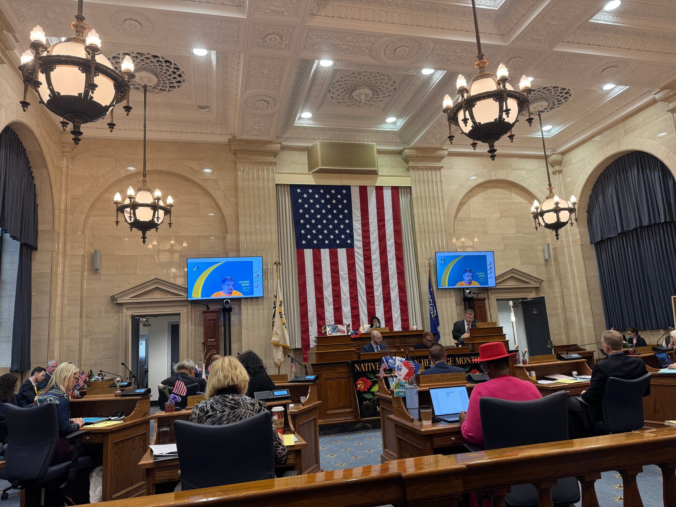 A legislative chamber with people seated at desks, a large American flag on the wall, chandeliers, and monitors displaying a speaker at the podium.