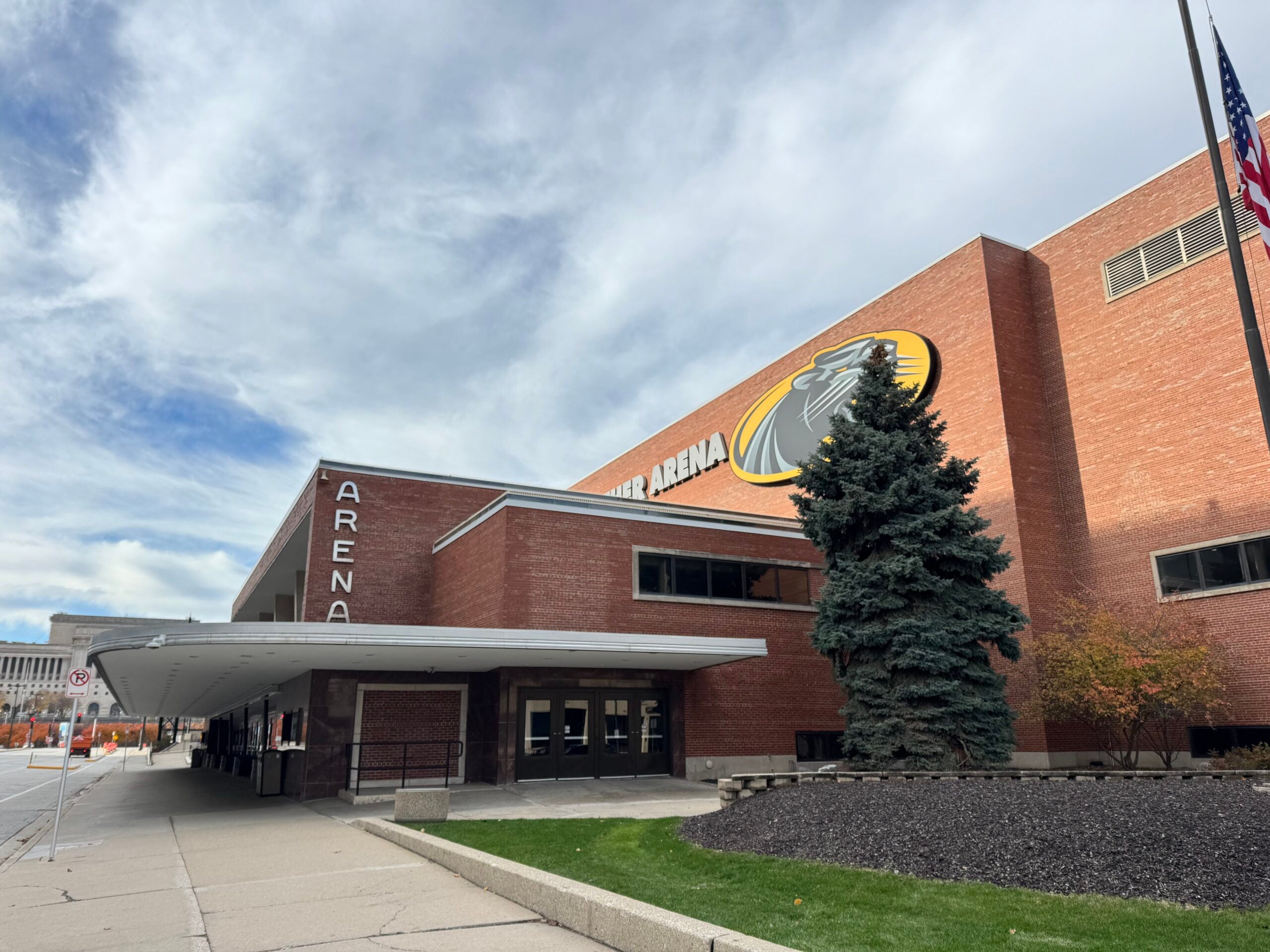 A brick arena building with large windows and a circular logo on the wall, an American flag, and a tree in front, under a partly cloudy sky.