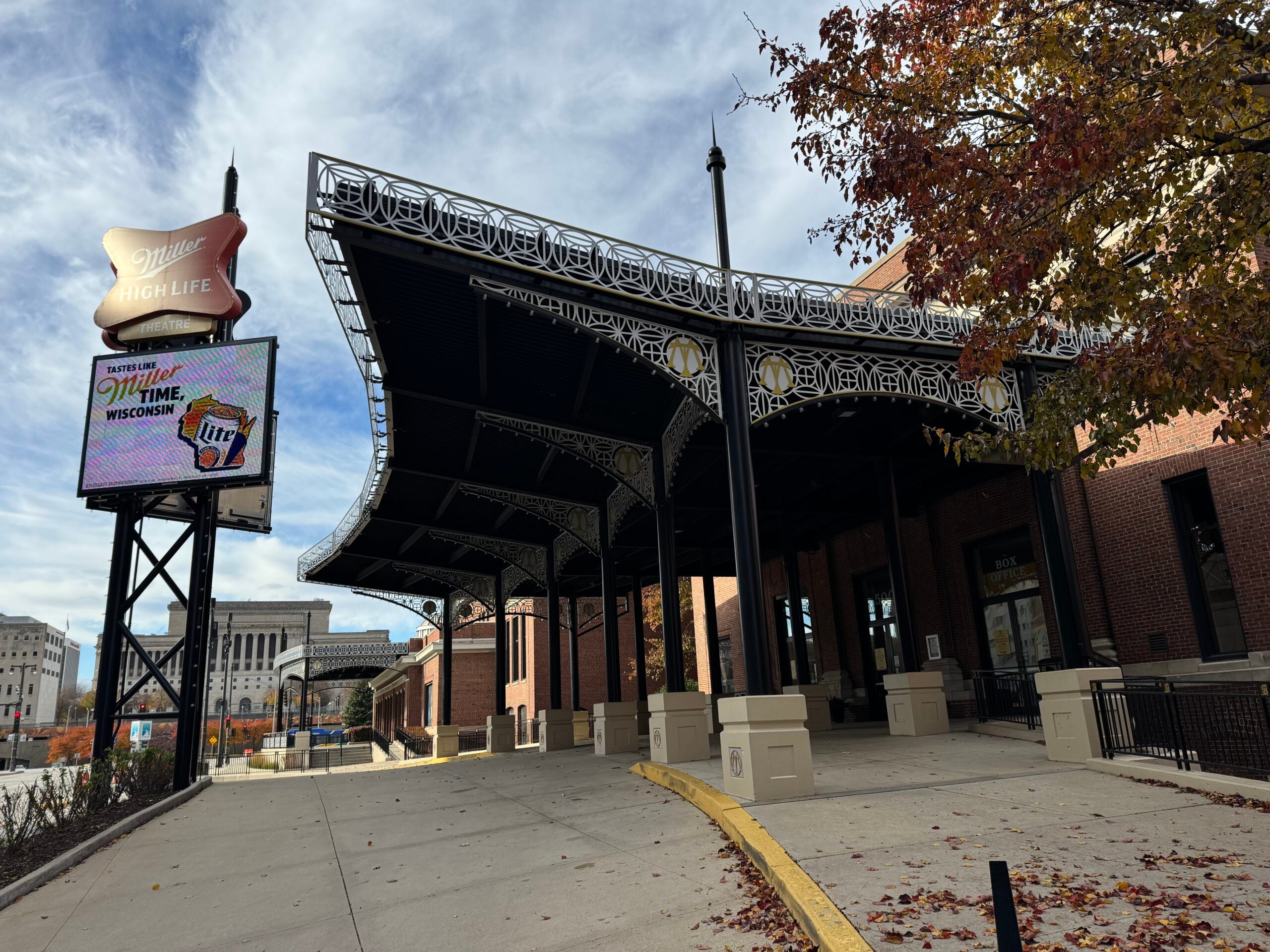 Covered outdoor entrance to a brick building with ornate black metalwork. A digital sign displays advertisements, and autumn leaves are scattered on the ground.