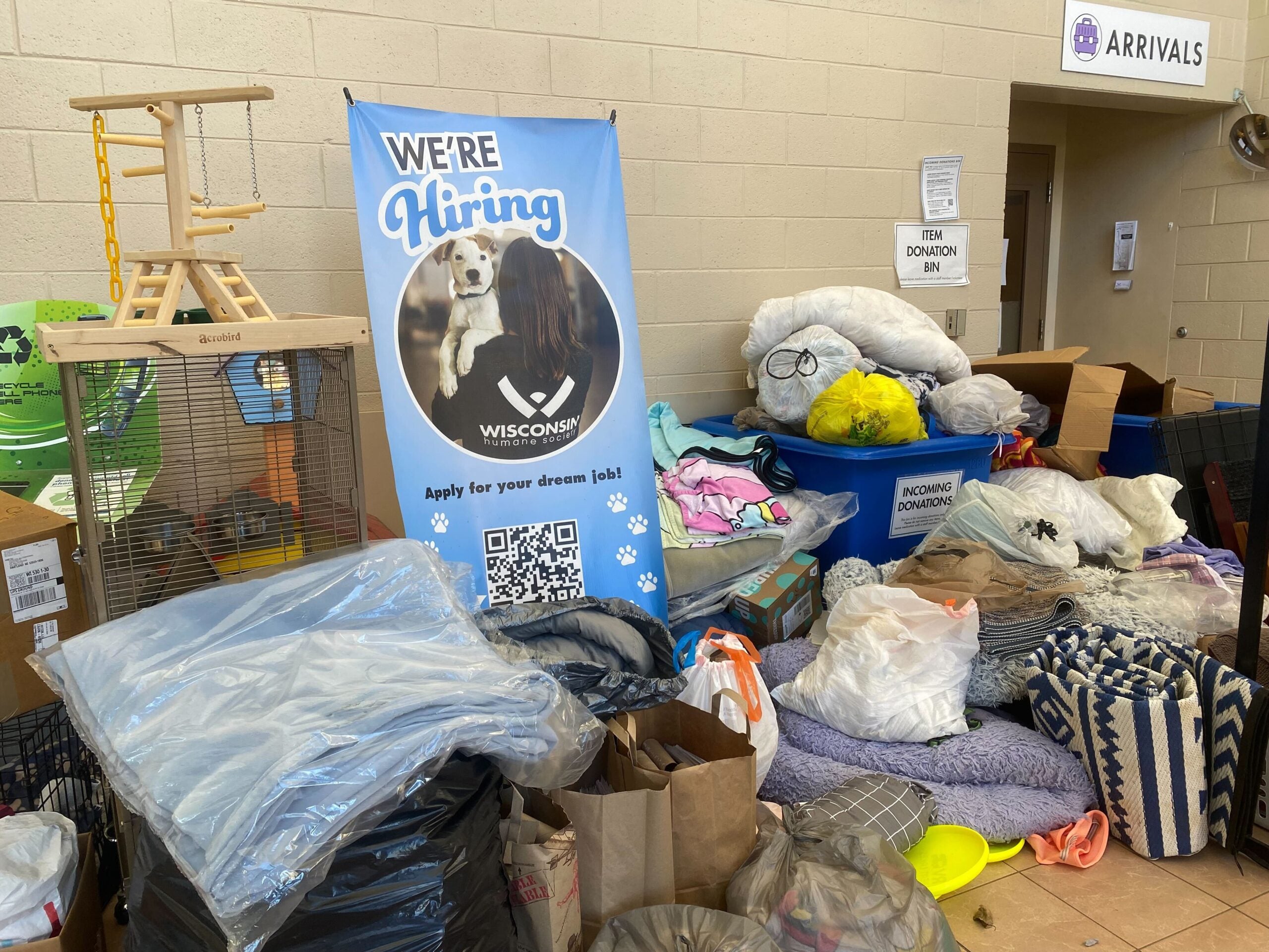 Piles of donated items and pet supplies near a Were Hiring banner at an animal shelter, with labeled bins and a birdcage visible in the background.