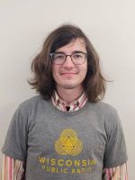 A person with long brown hair and glasses wears a gray Wisconsin Public Radio T-shirt over a striped collared shirt, standing in front of a plain light-colored wall.