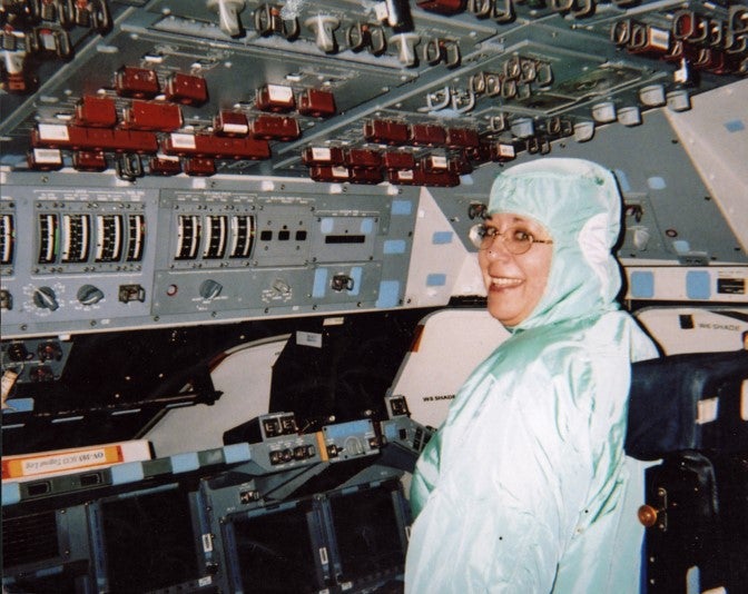 A person in a protective suit sits at the controls of a spacecraft cockpit, smiling at the camera.