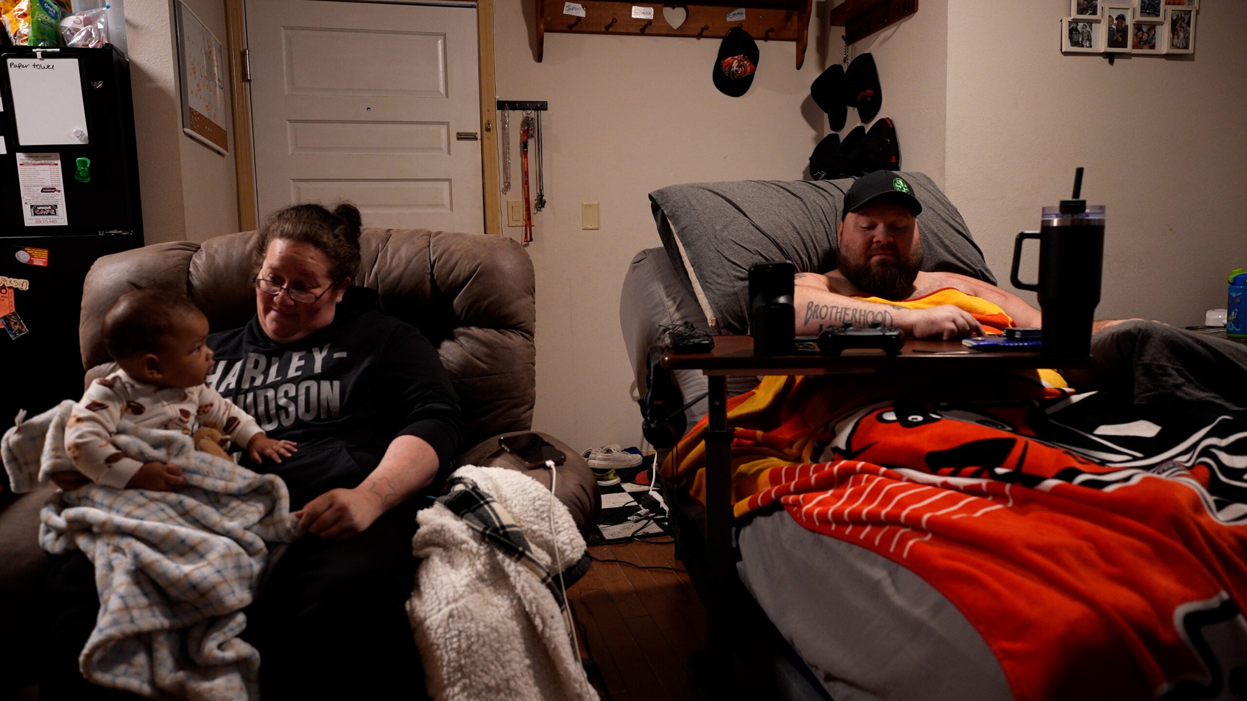 A woman holds a baby in an armchair while a man sits in a reclining bed using a laptop; both are in a living room with personal items and blankets.
