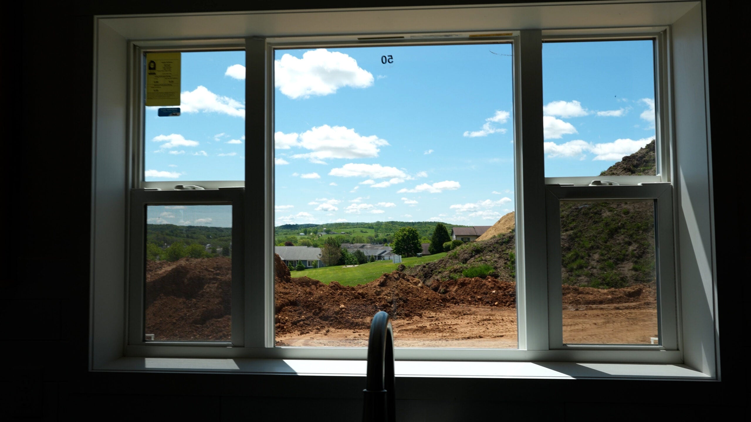 A view through a kitchen window showing a construction site with dirt piles, green hills, houses, and a blue sky with clouds in the background.