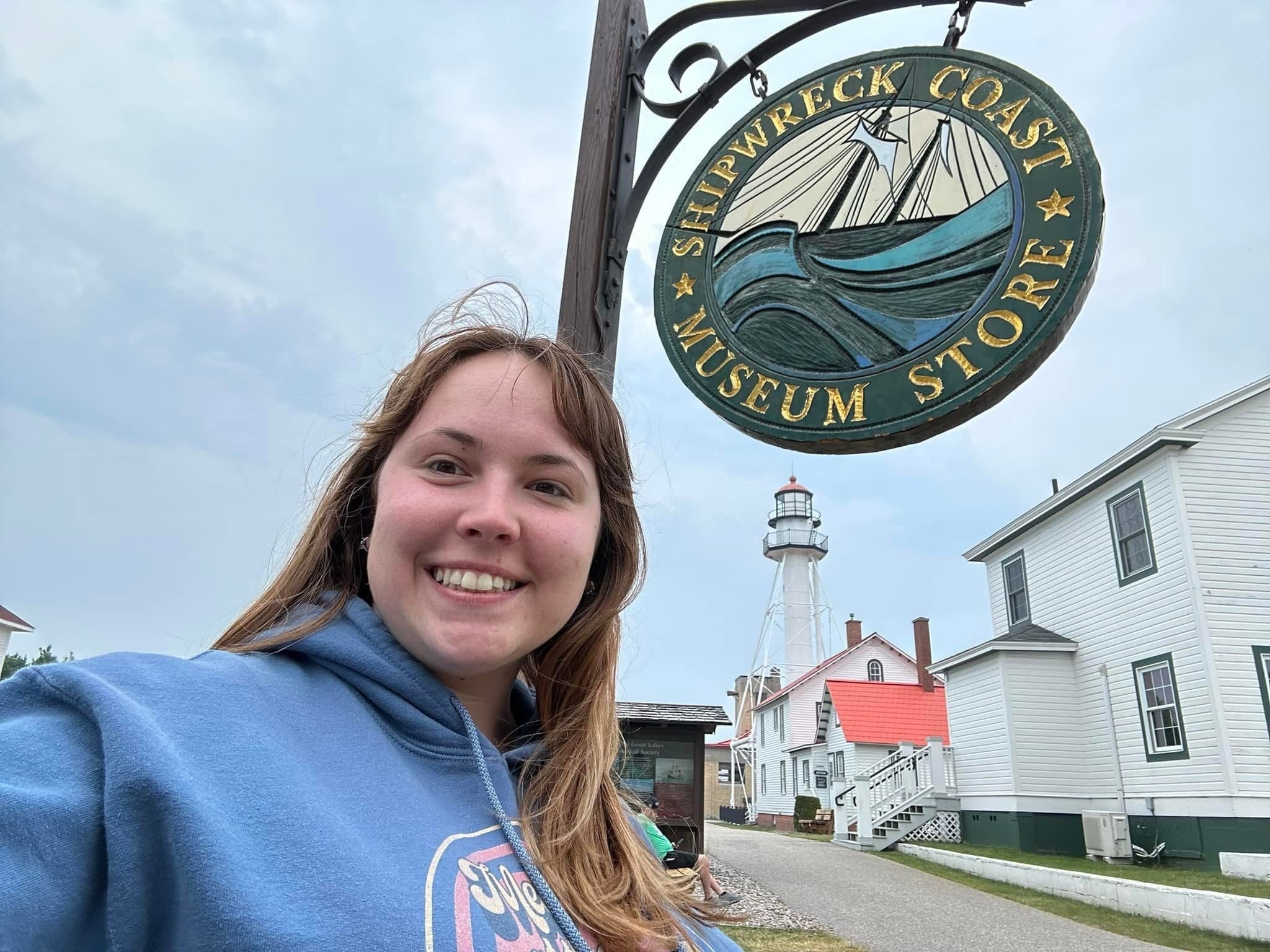 A person in a blue hoodie smiles in front of a Shipwreck Coast Museum Store sign, with a lighthouse and white buildings visible in the background.