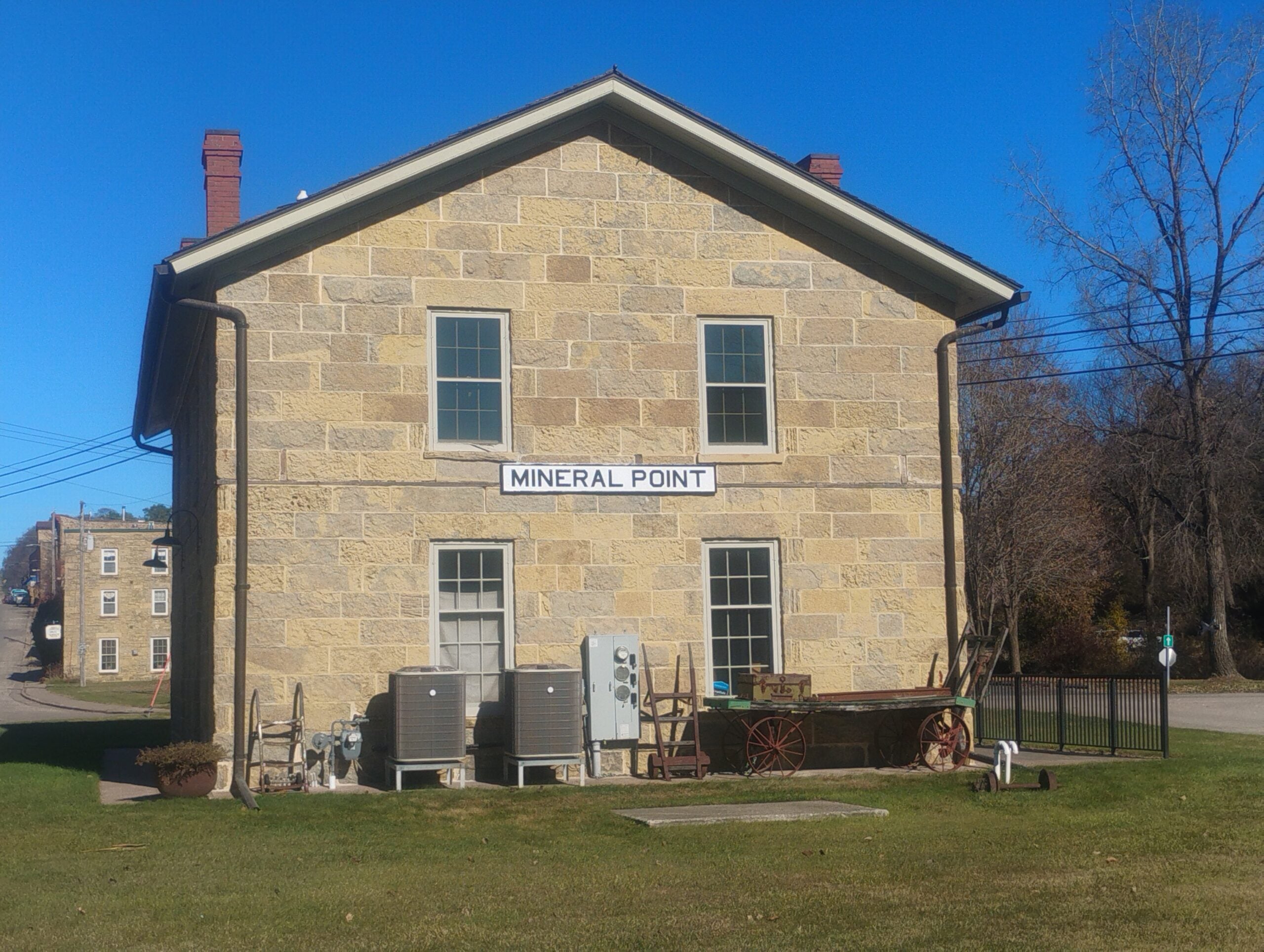Stone building labeled Mineral Point with two chimneys, several windows, outdoor HVAC units, and old farm equipment on a grassy lawn under a clear blue sky.