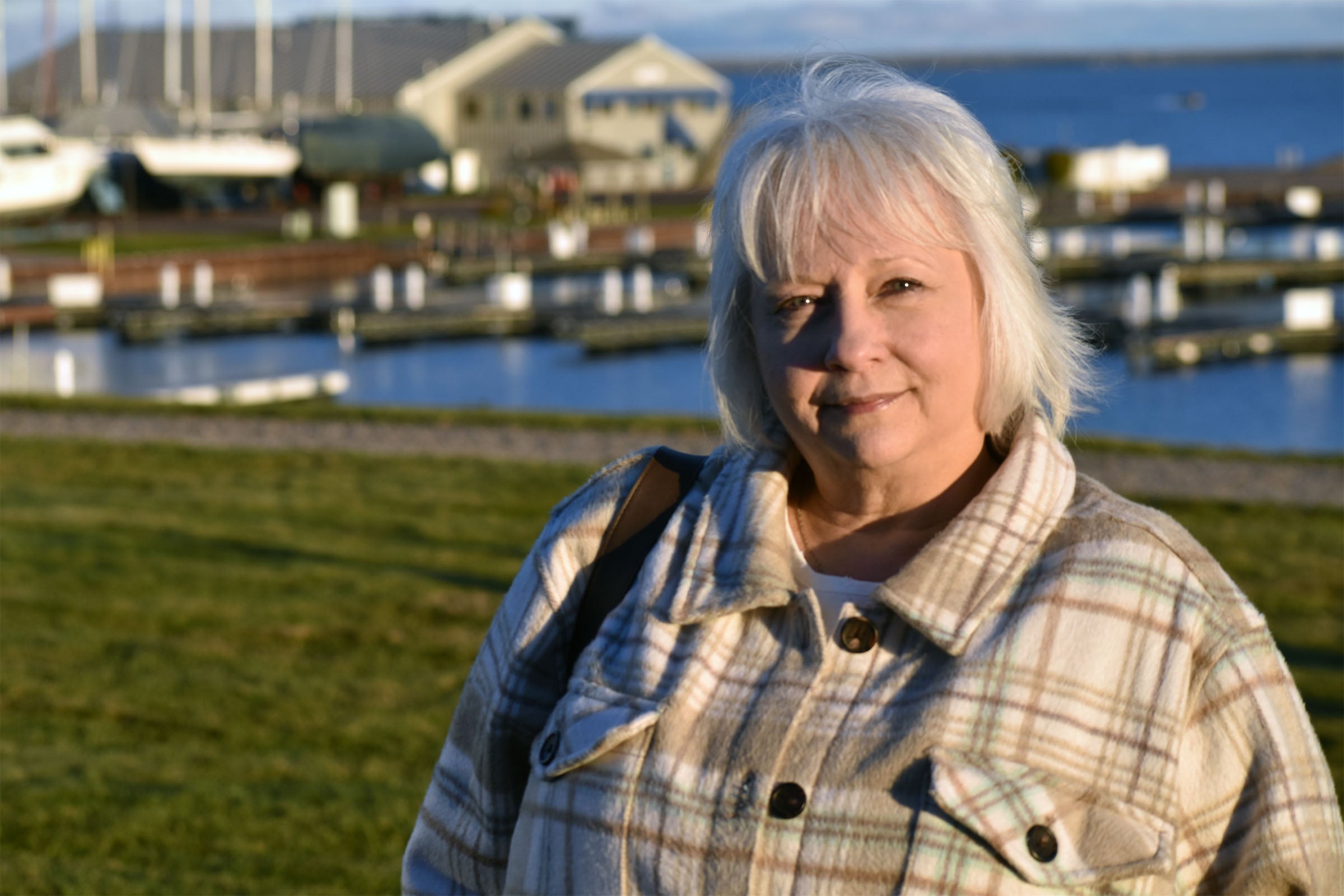 Woman with gray hair wearing a plaid jacket stands outdoors near a marina with boats and buildings in the background.