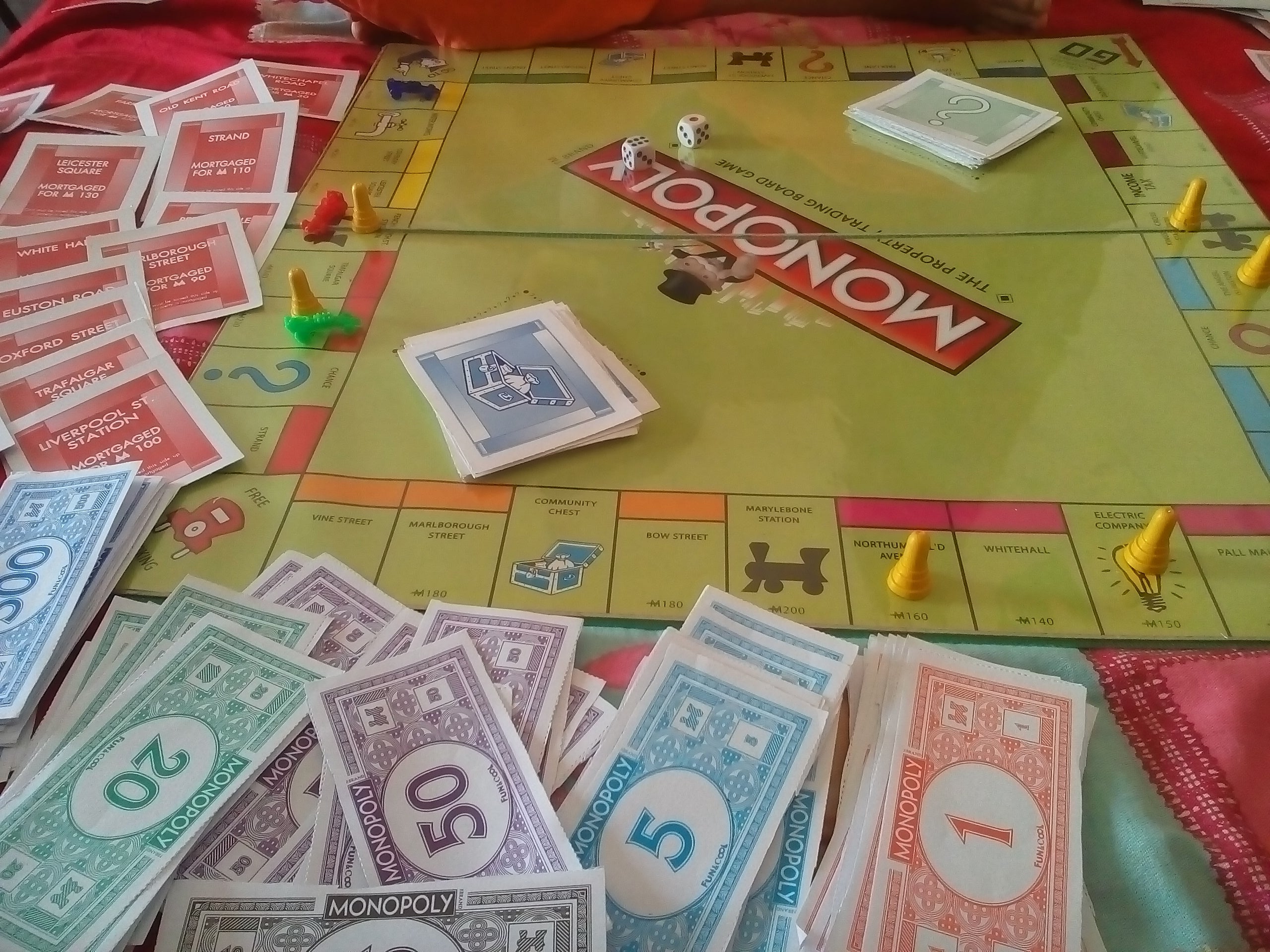 A game of Monopoly is in progress, showing the board, cards, dice, player tokens, and various denominations of Monopoly money.