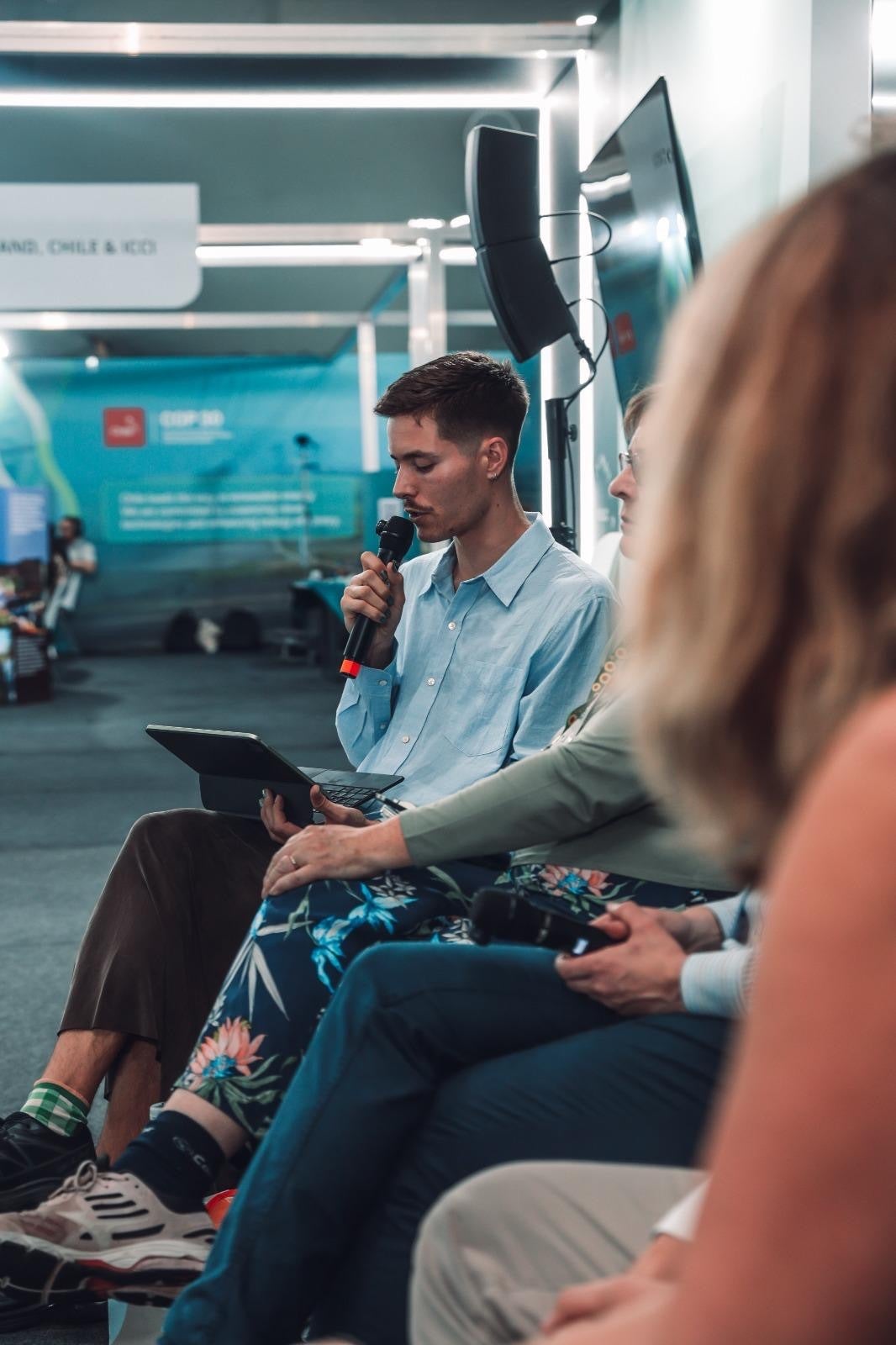 A man sits on a panel holding a microphone and tablet, speaking to an audience. Other panelists are seated beside him in a modern indoor setting.