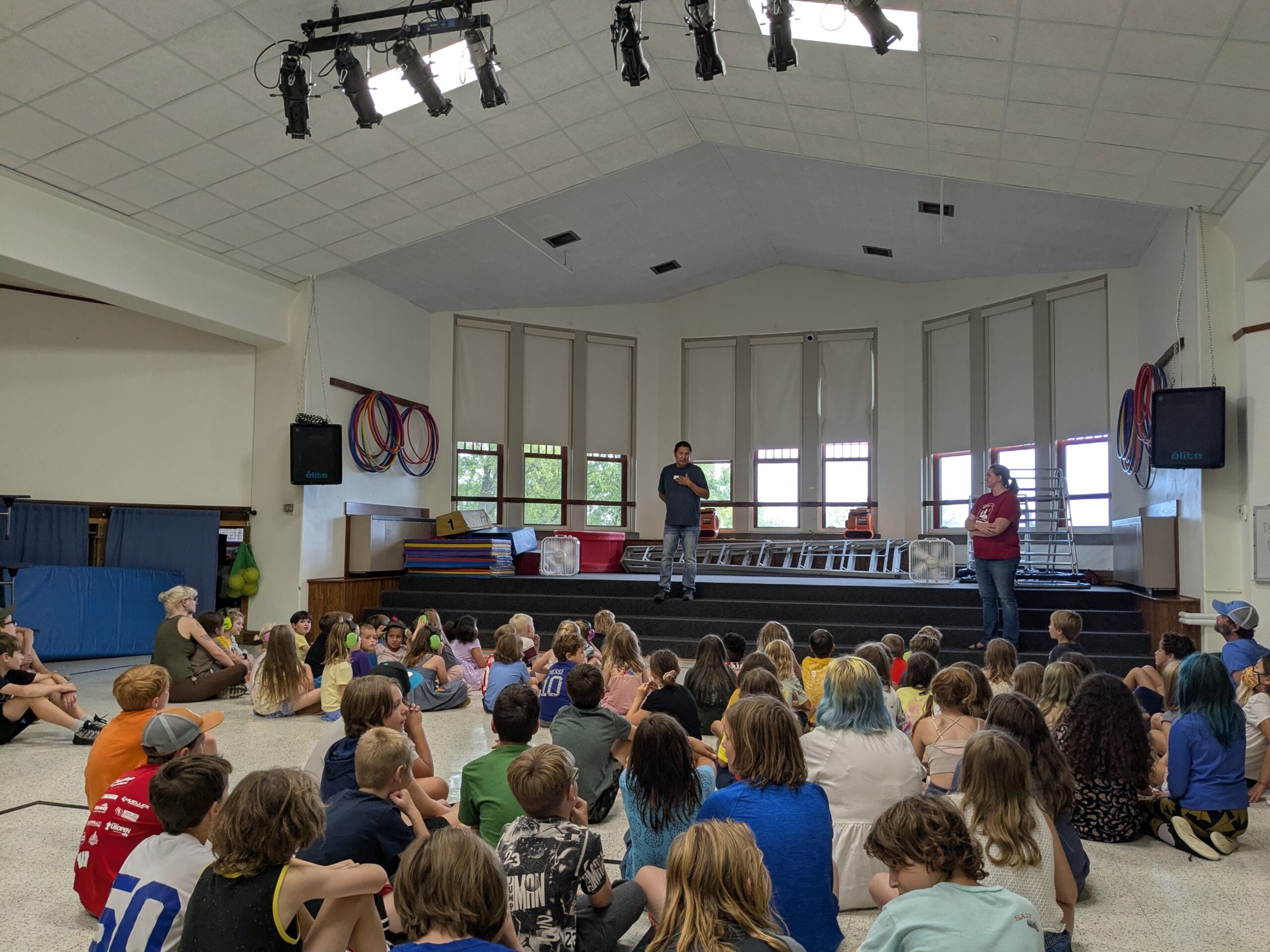 A group of children sit on the floor facing a stage where two adults are speaking in a gymnasium-like room with high ceilings and large windows.