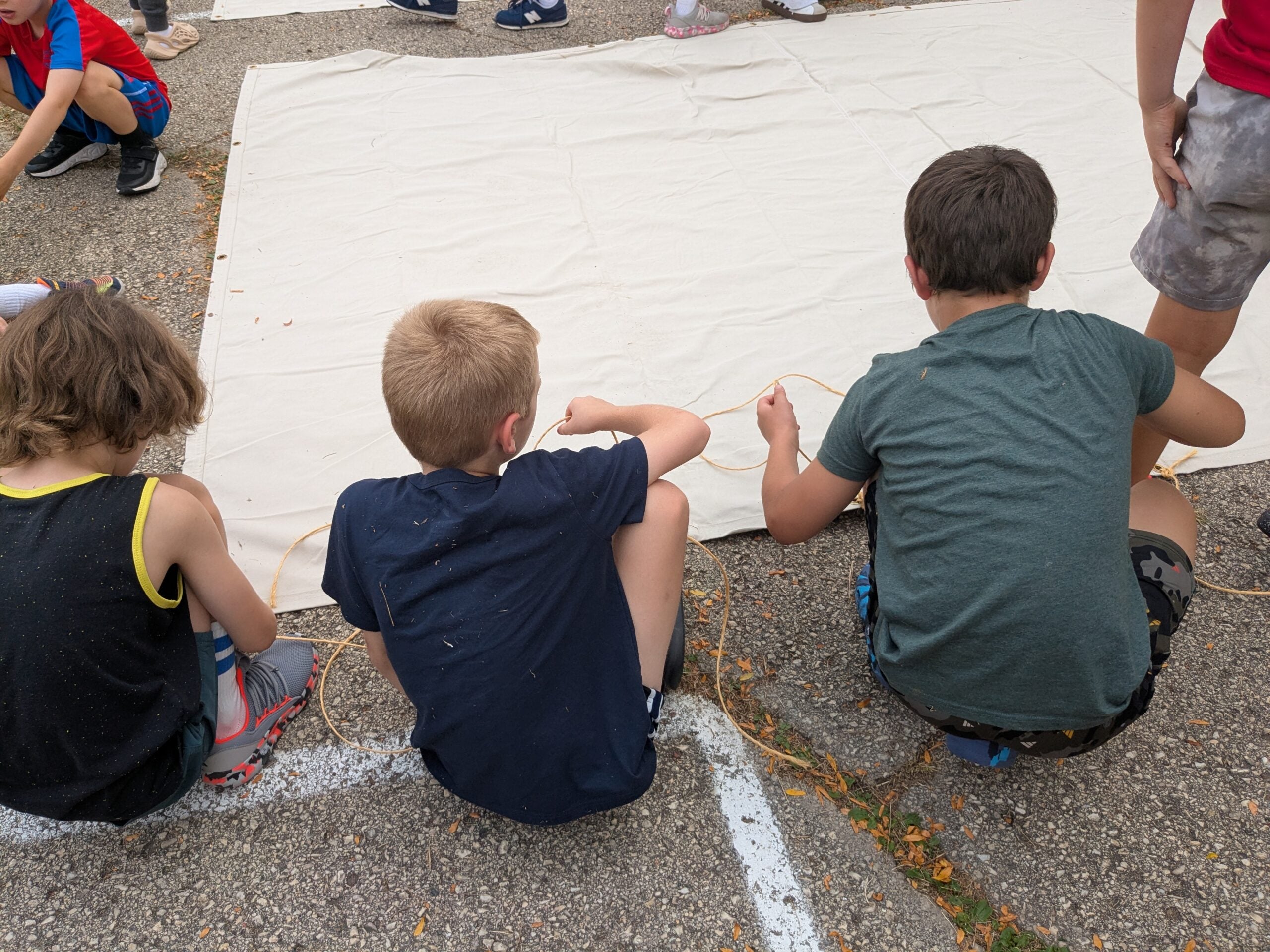 Three children sitting on the ground work with ropes near a large white canvas or tarp during an outdoor activity.