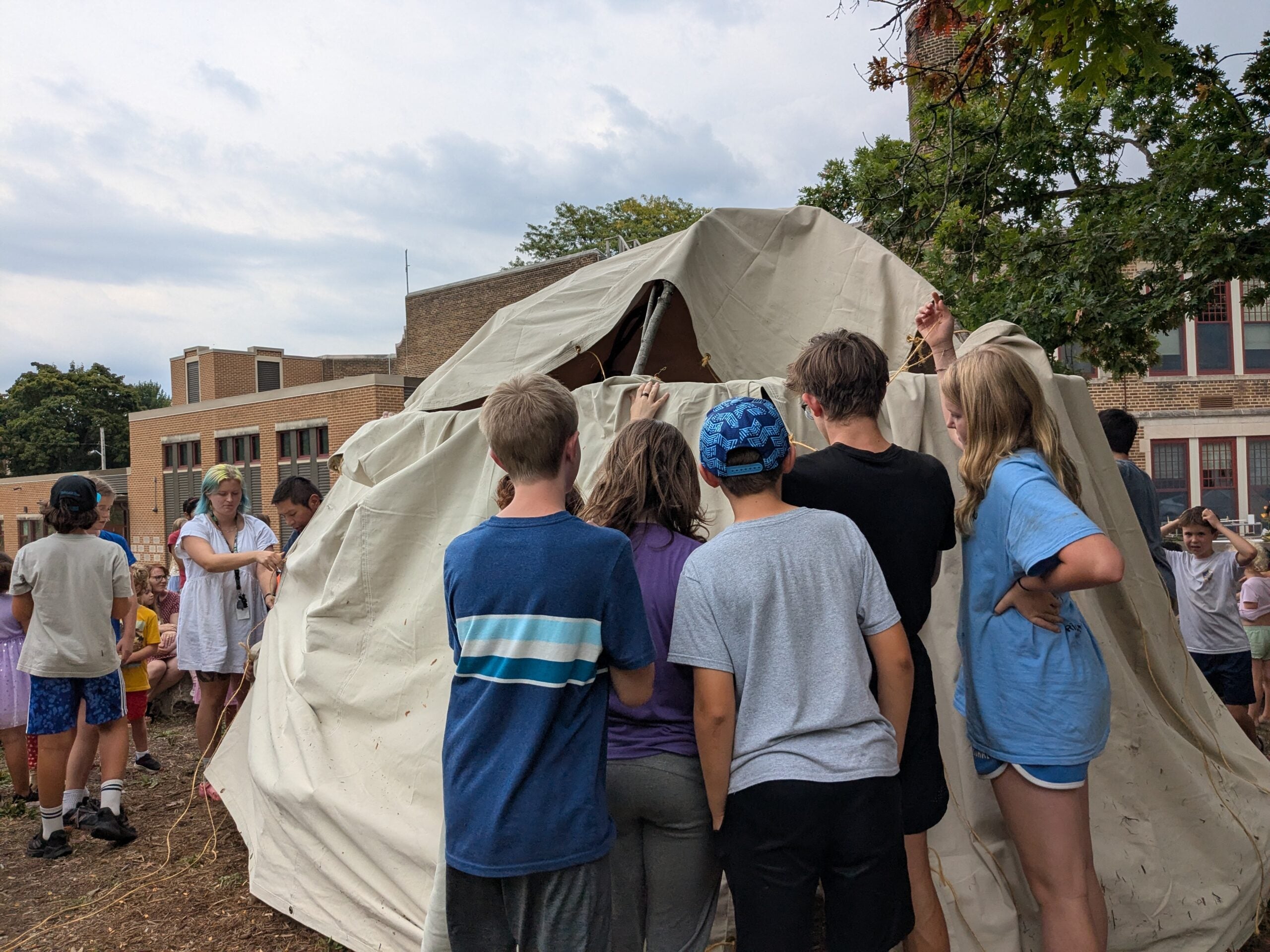 A group of children stand around and look inside a large canvas tent outdoors near a school building on a cloudy day.