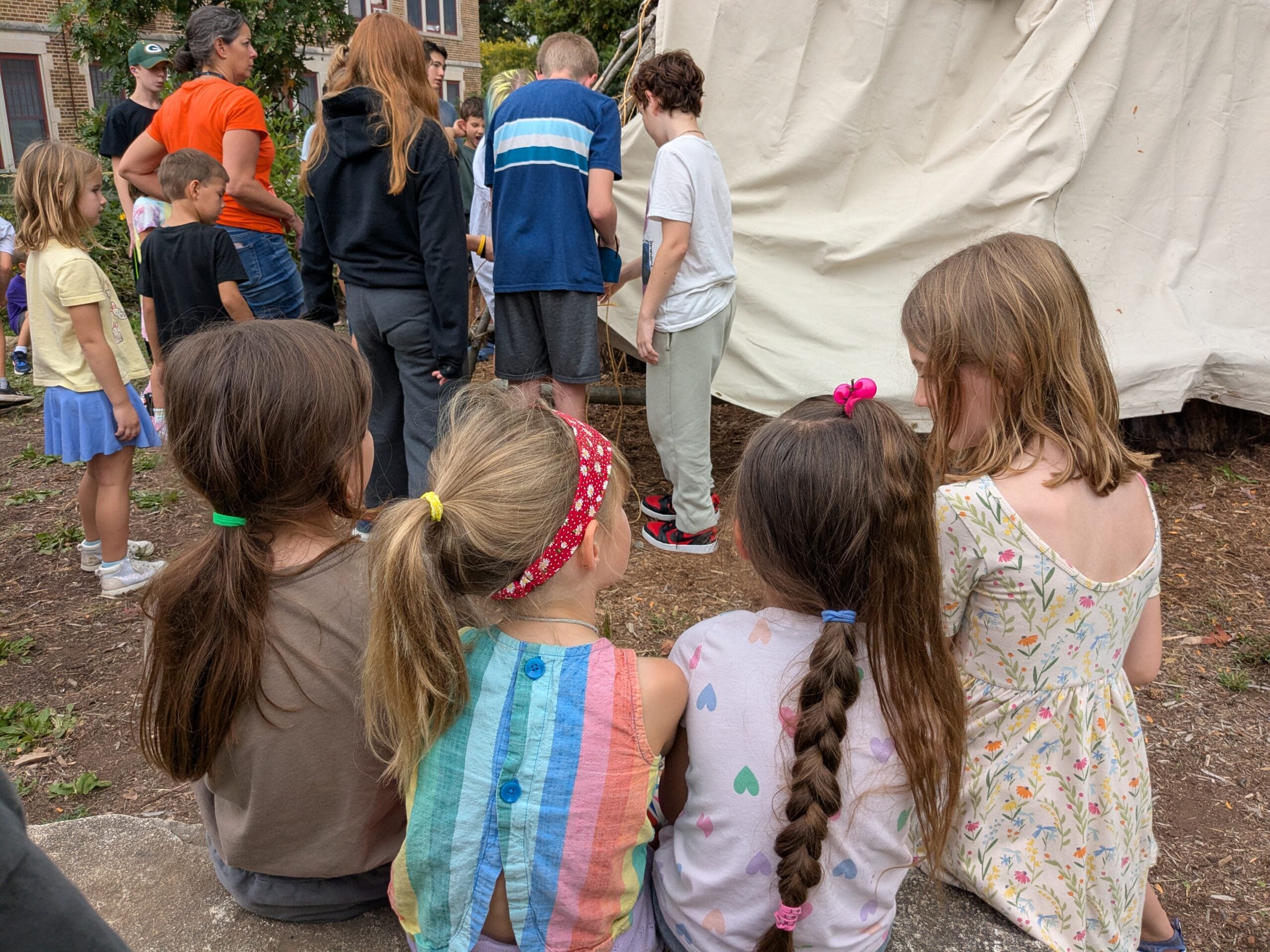 A group of children sit on a stone ledge, watching other kids and adults gathered near a large white canvas tent outdoors.