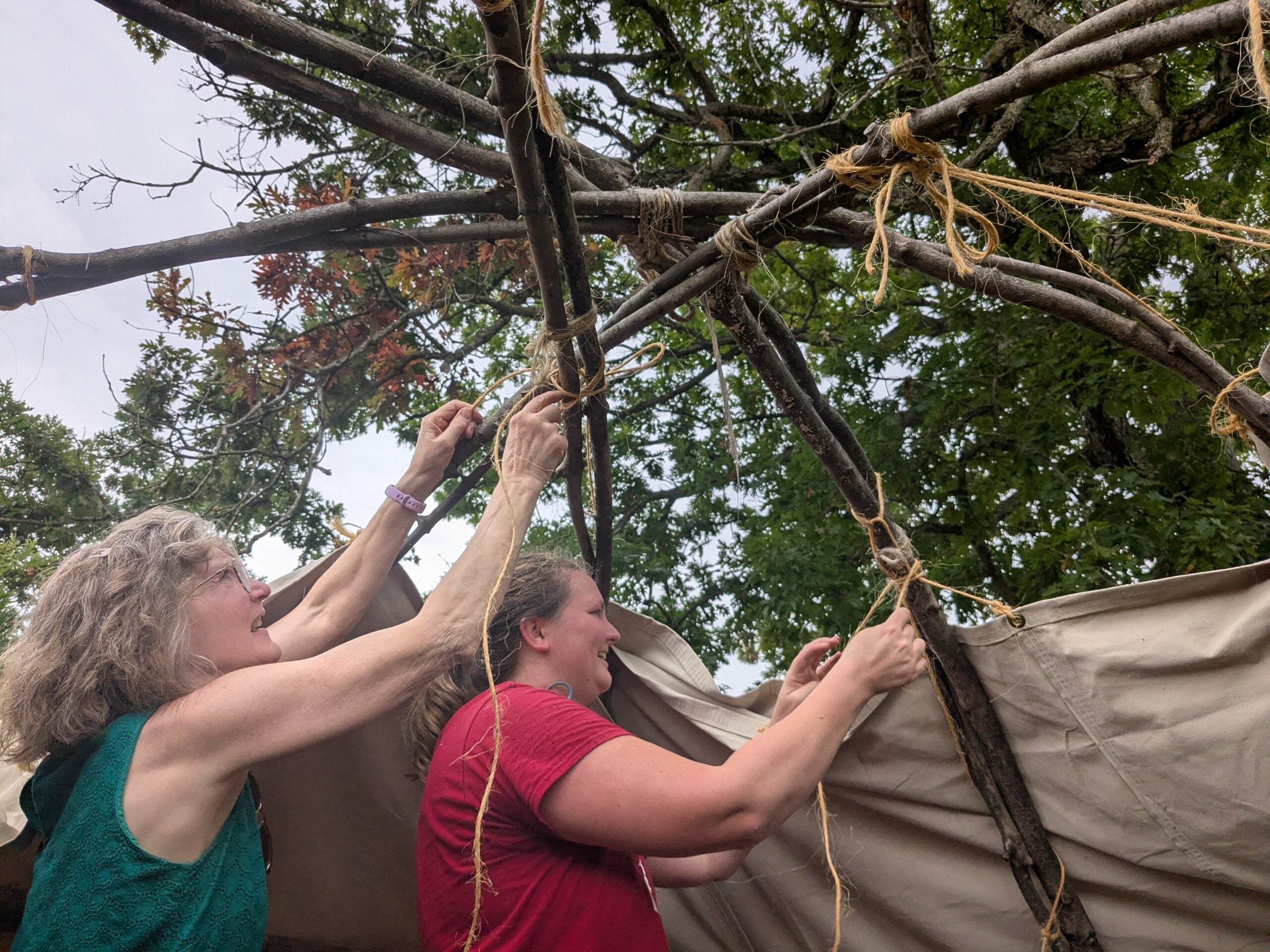 Two women tie tree branches together with rope, constructing a wooden frame outdoors under leafy trees.