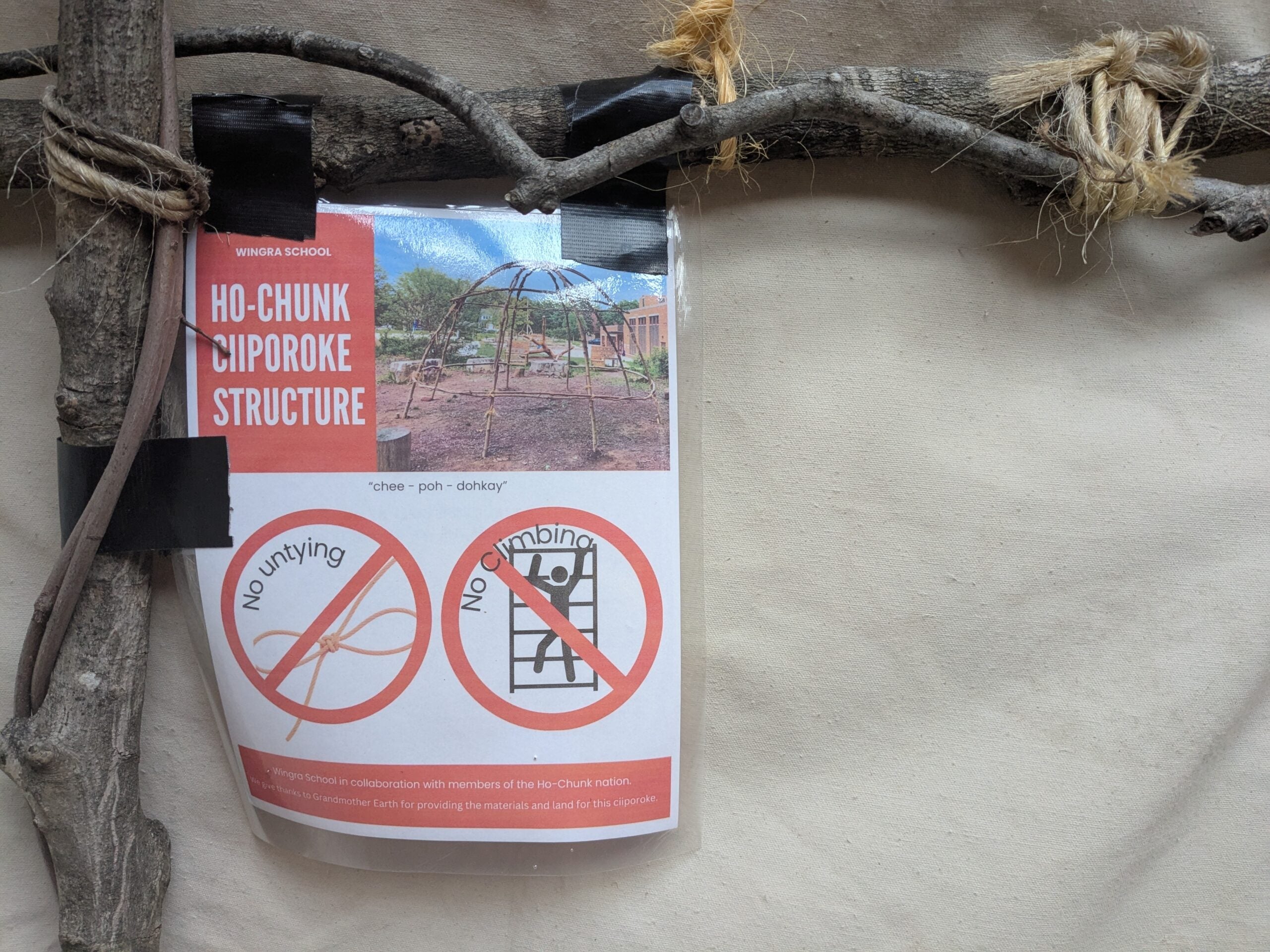 A sign attached to wooden branches reads Ho-Chunk Ciiporoke Structure and displays icons for No Untying and No Climbing with explanatory text.