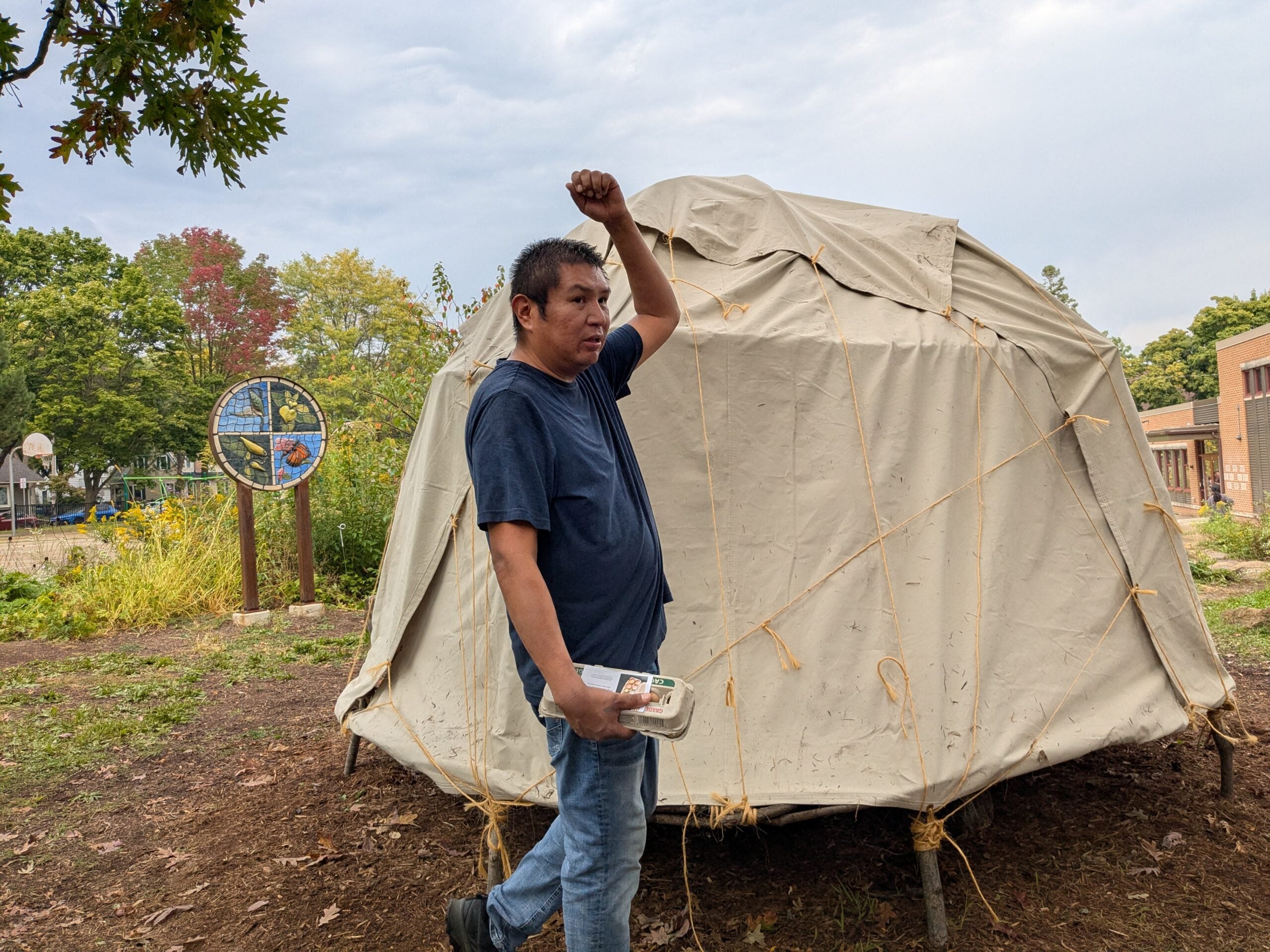 A man stands outdoors holding a package and raising one arm in front of a covered dome structure, with trees and a circular sign in the background.