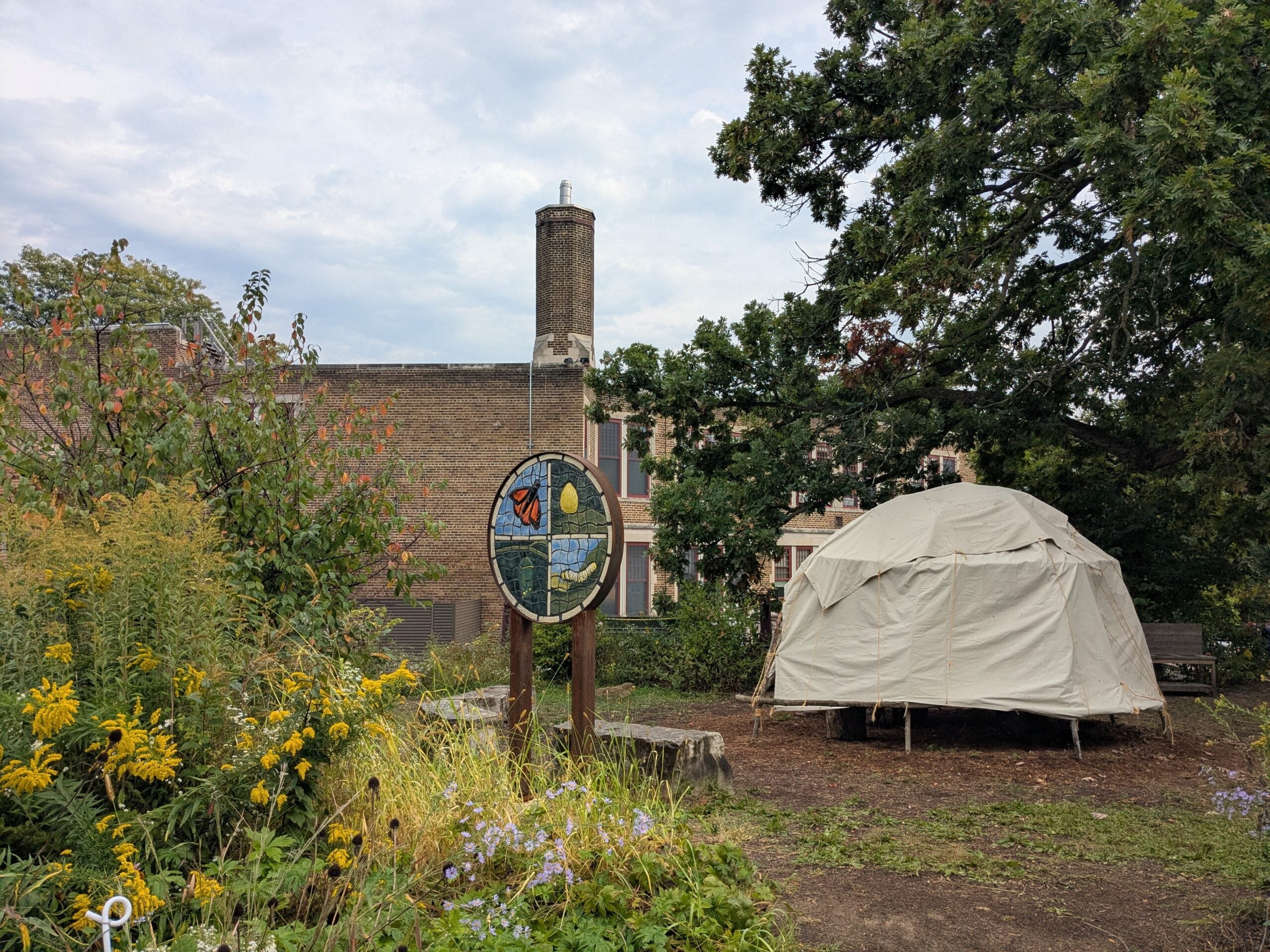 A white canvas tent stands beside a colorful stained glass sign in a garden with wildflowers and trees, near a brick building.