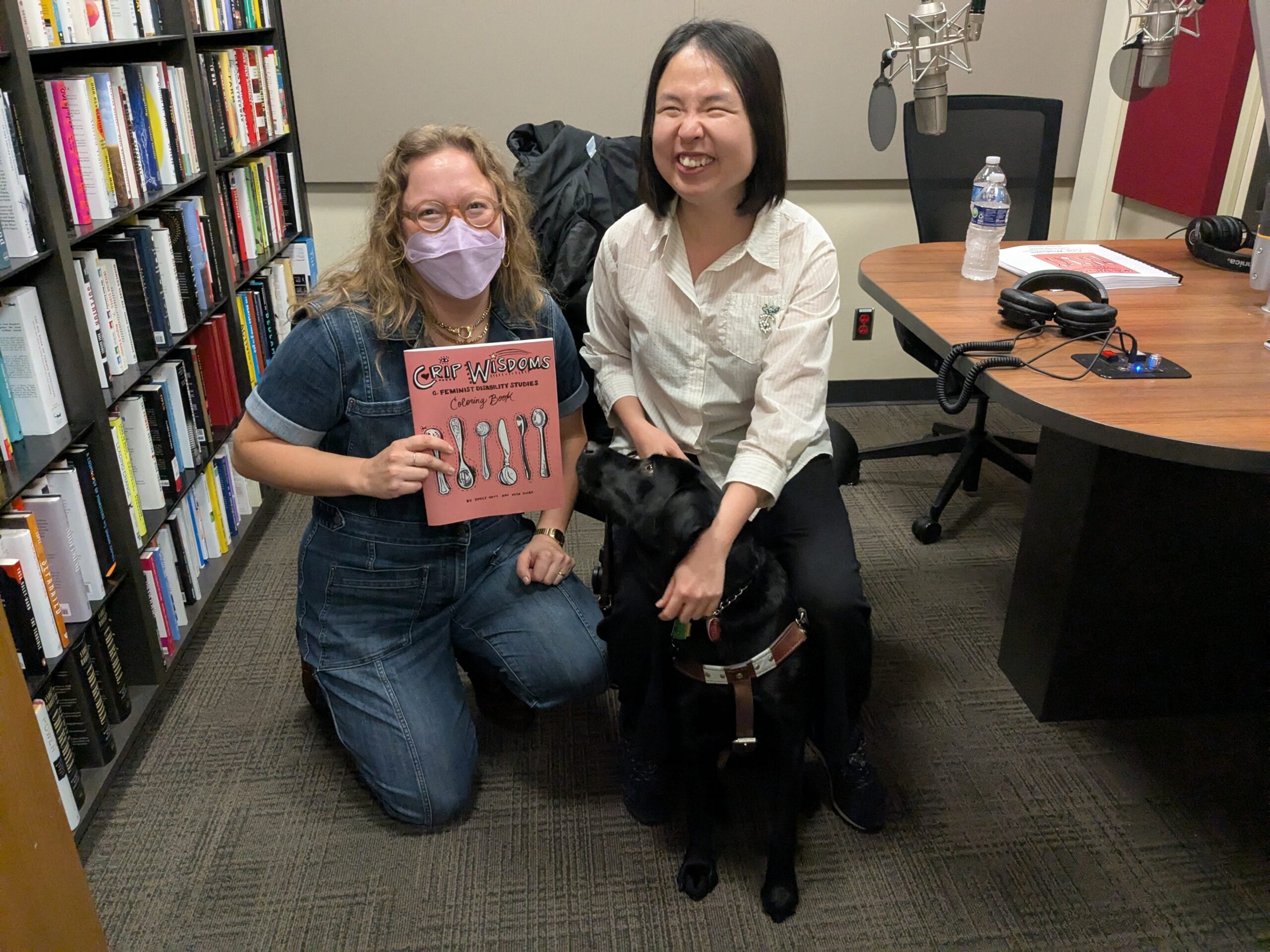 Two people pose in a recording studio. On the left, a person in a light purple mask holds a pink book titled "Crip Wisdoms." On the right, a person in a white shirt smiles. A black guide dog is sitting between them.