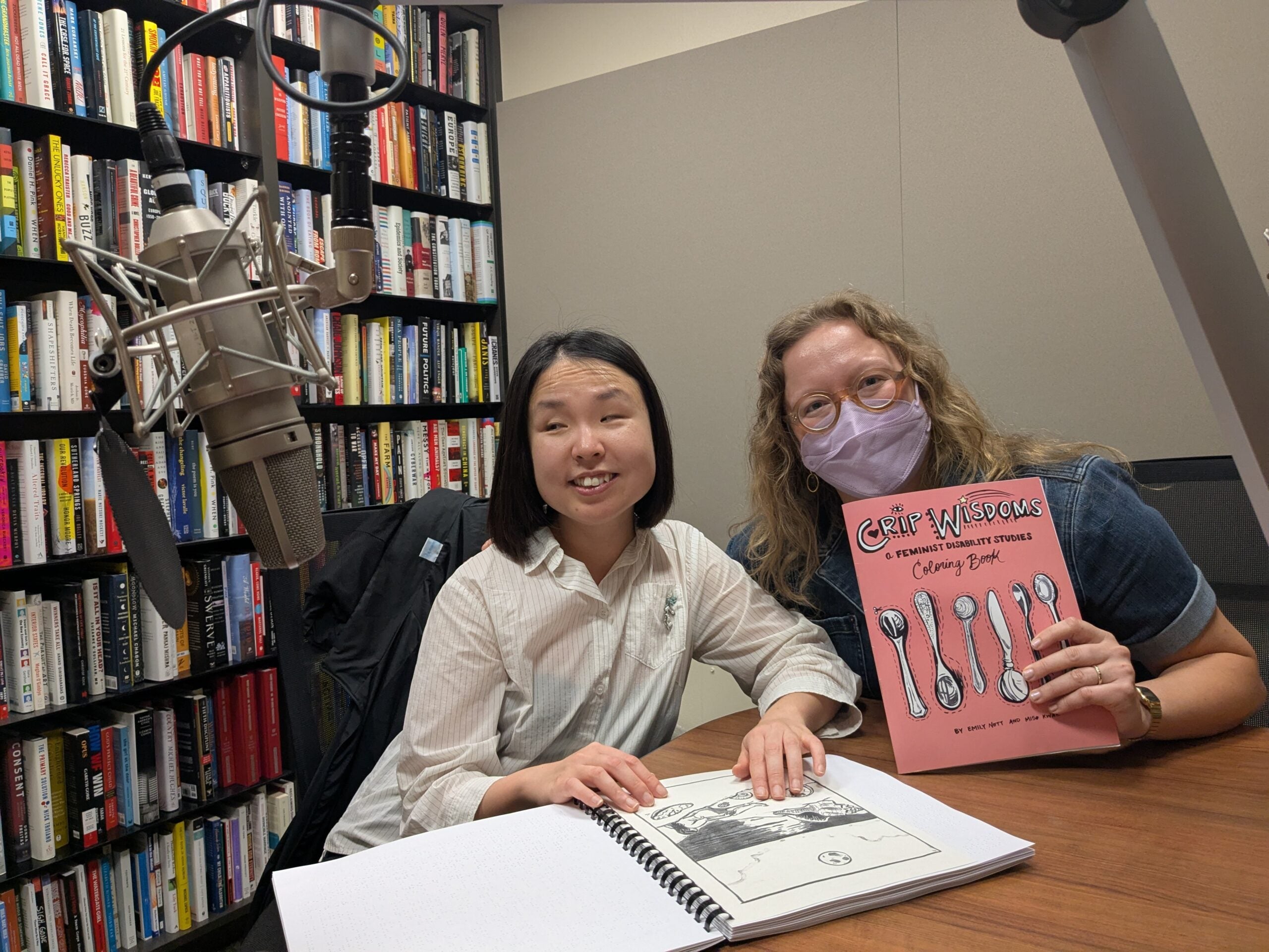 Two women sit at a table in a library or office, one holding a cookbook titled Chef Wisdom, while the other has an open Braille book; a microphone is visible on the left.