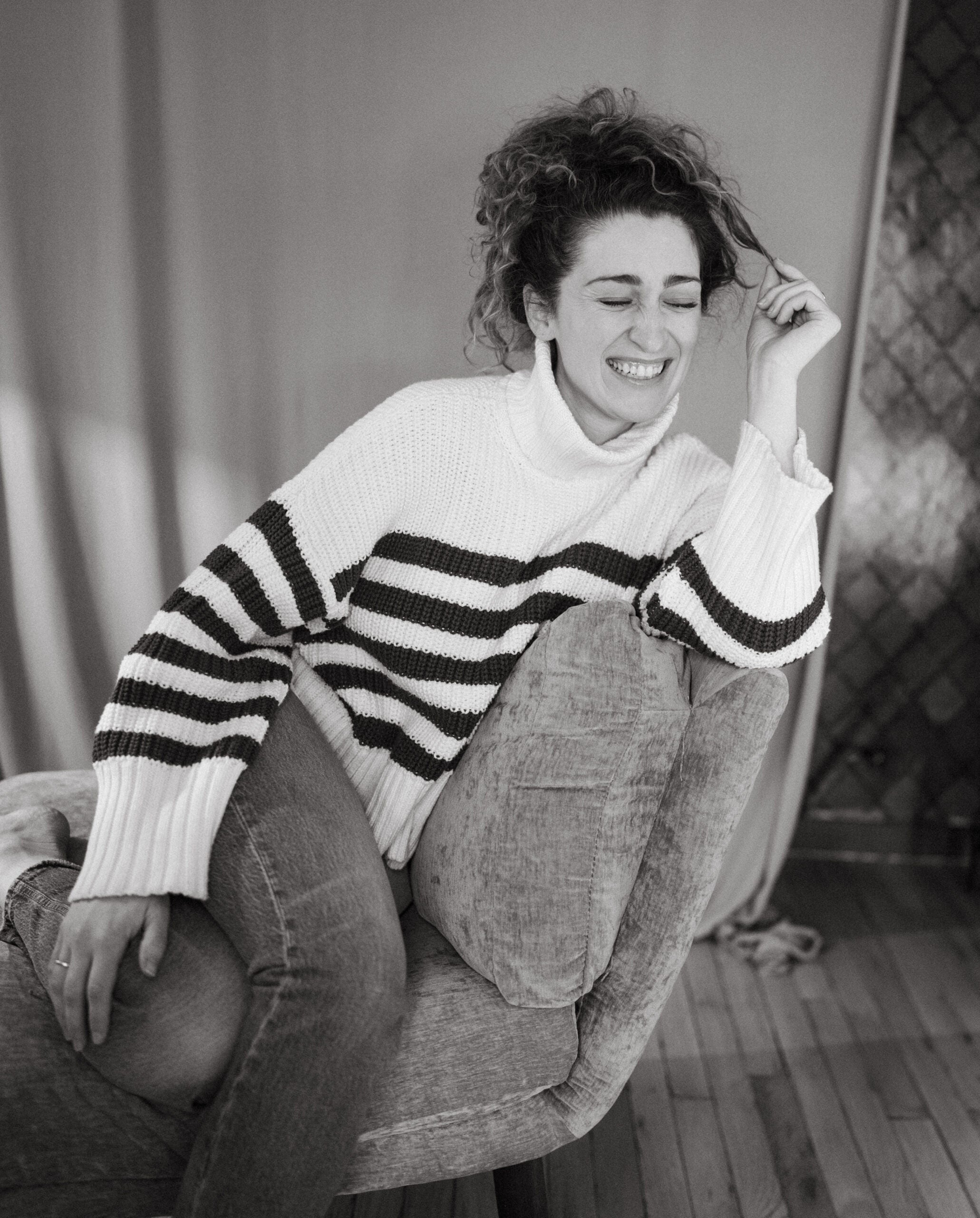 A person with curly hair sits sideways on a cushioned chair, wearing a striped sweater and jeans, smiling with eyes closed in a relaxed indoor setting.