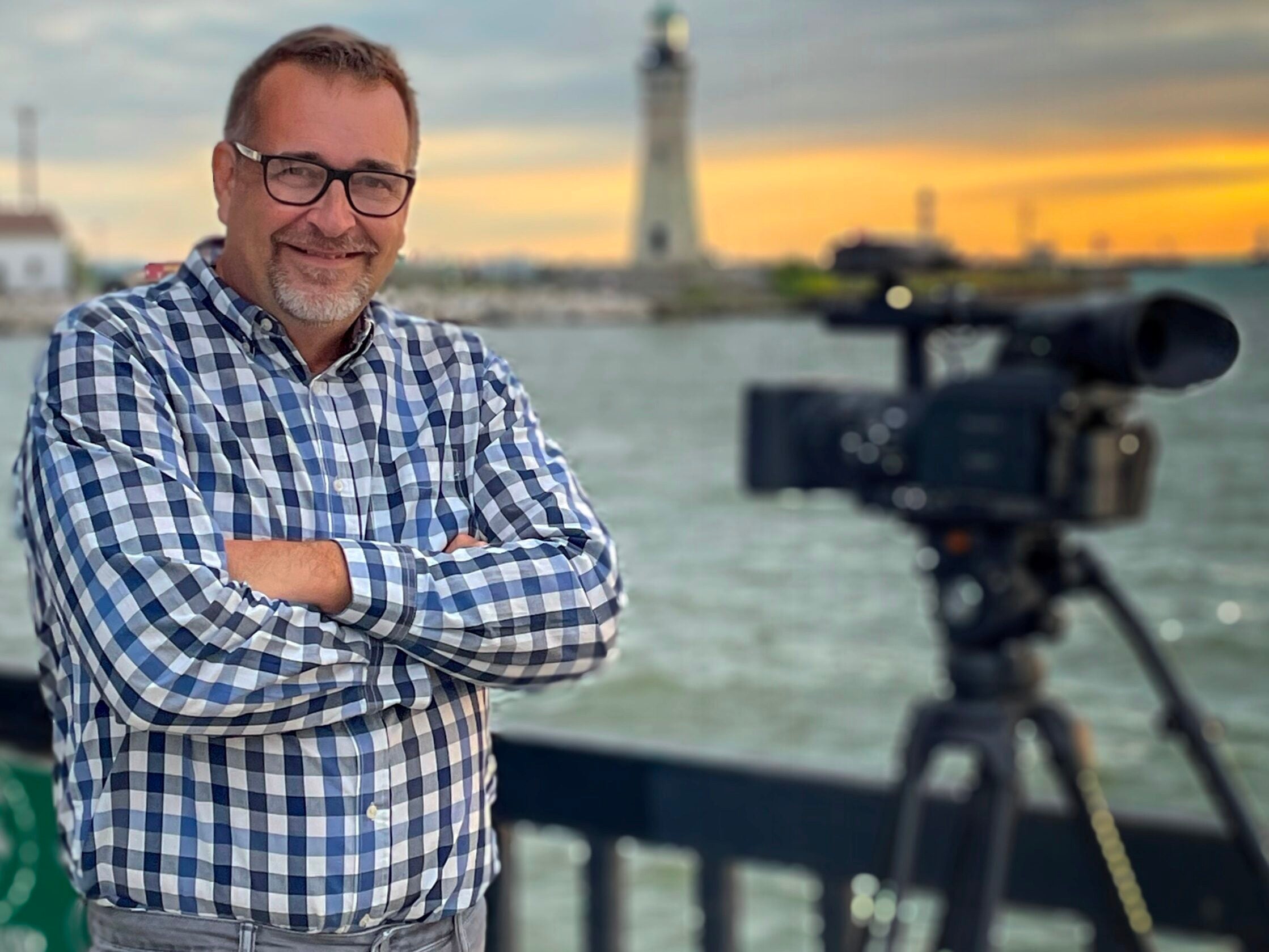 A man in a plaid shirt stands smiling with arms crossed by a waterfront, next to a video camera on a tripod, with a lighthouse in the background at sunset.