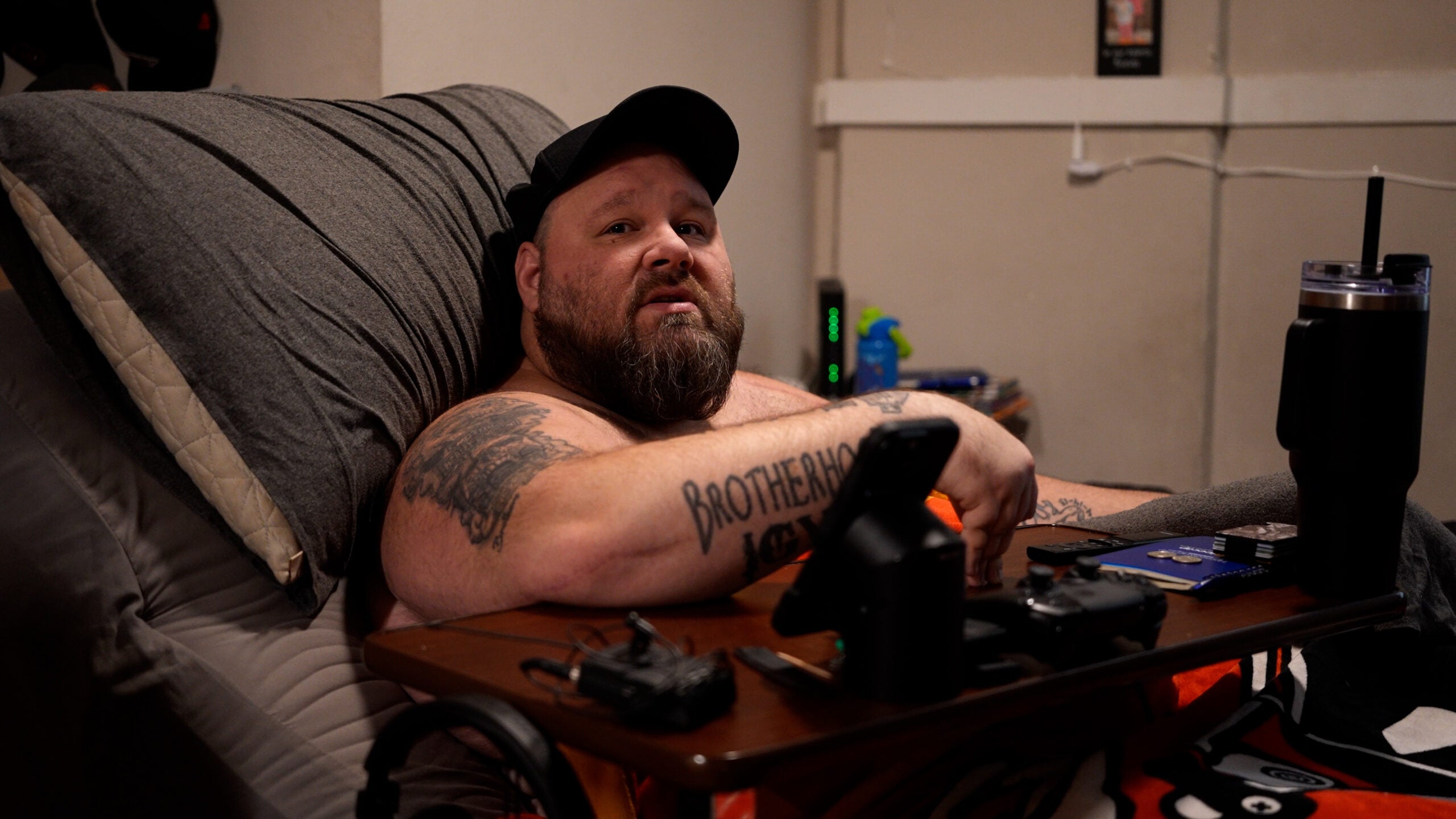 A bearded man with tattoos reclines in bed, wearing a black cap. A tray with electronic devices, a drink, and a controller rests on his lap.