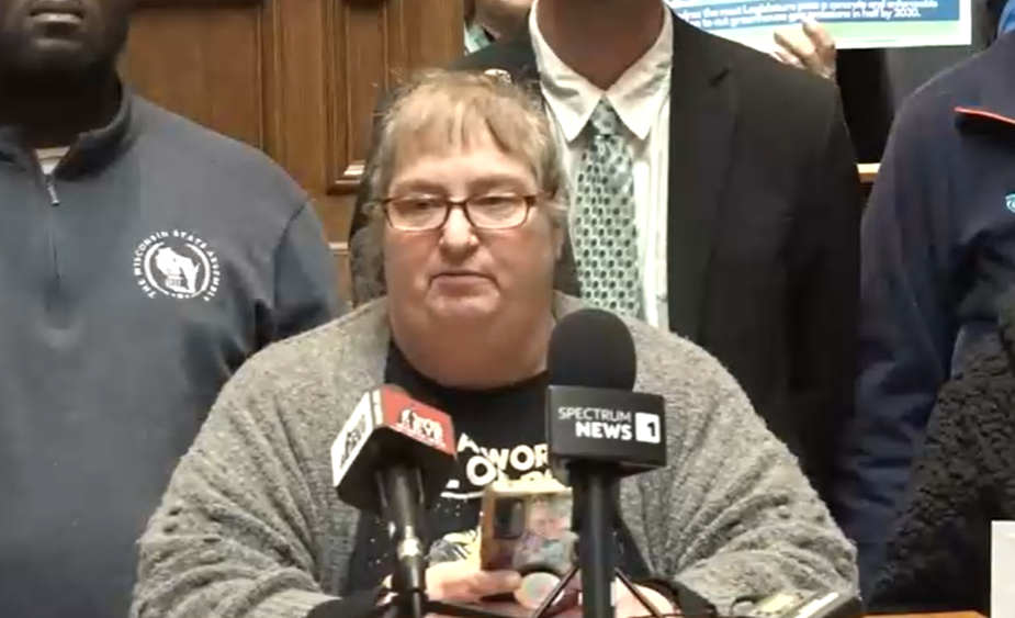 A woman speaks at a podium with multiple microphones during a press event, with several people standing behind her in a wood-paneled room.