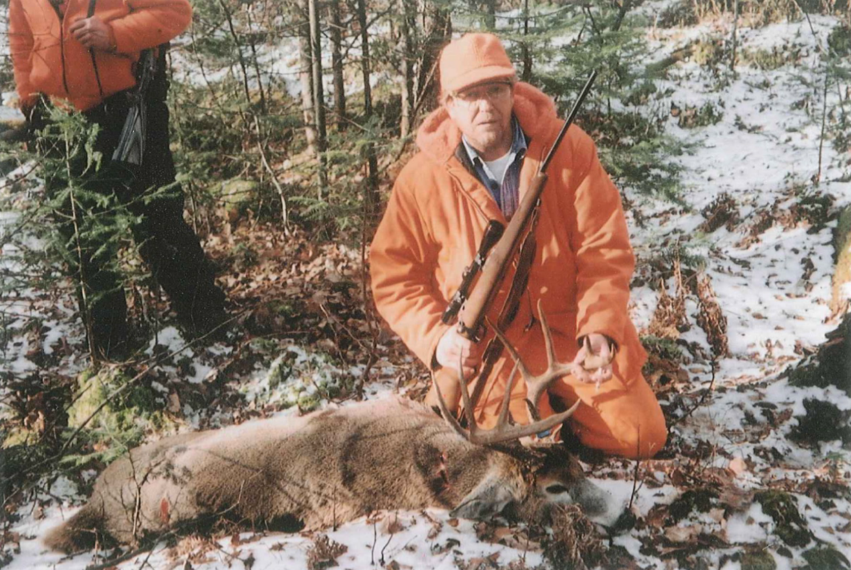 A person in orange hunting gear kneels next to a deer with antlers on a snowy forest floor, holding a rifle.