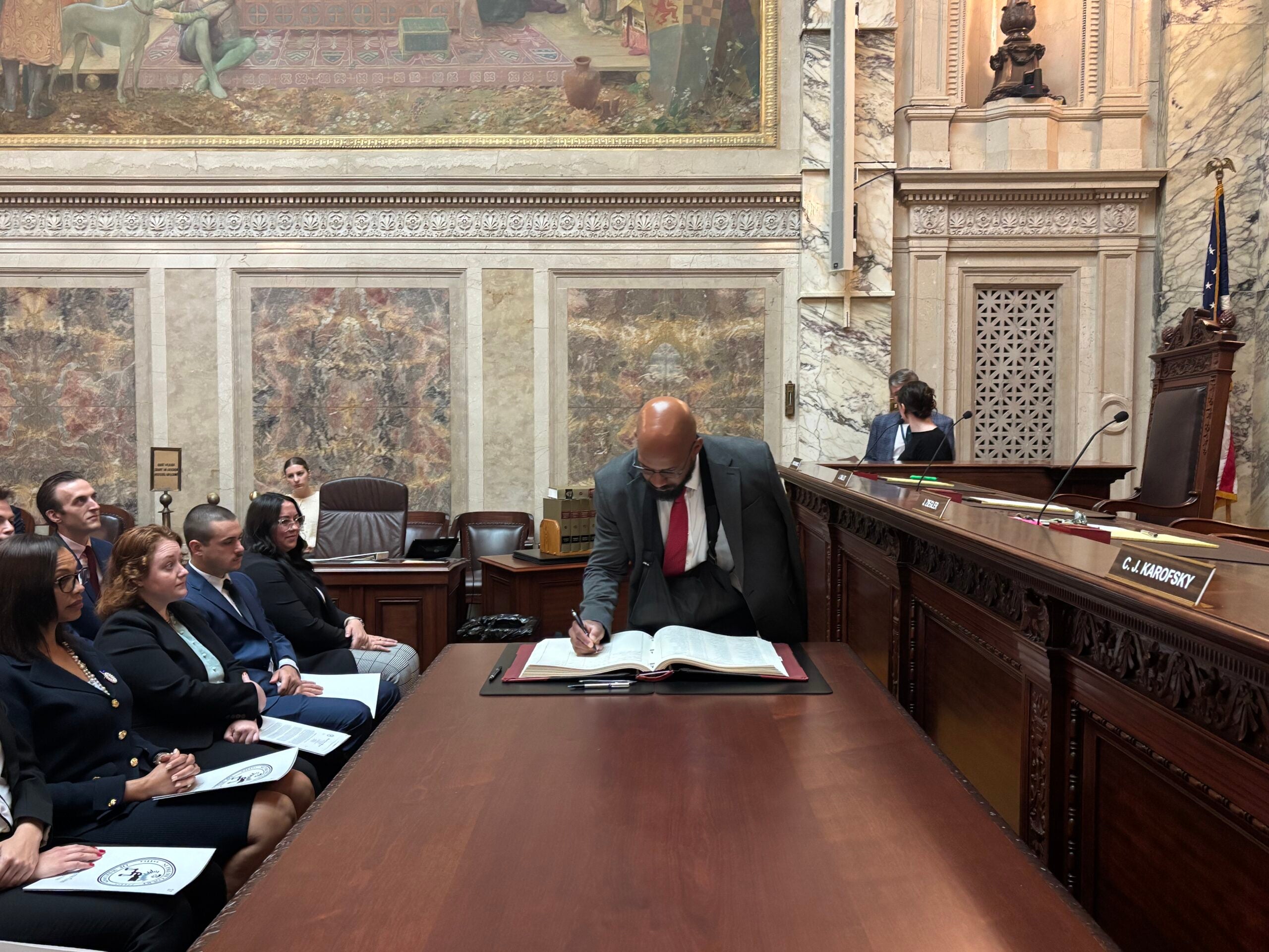 A man in a suit signs a large book at the front of a courtroom as several people seated to the side watch.